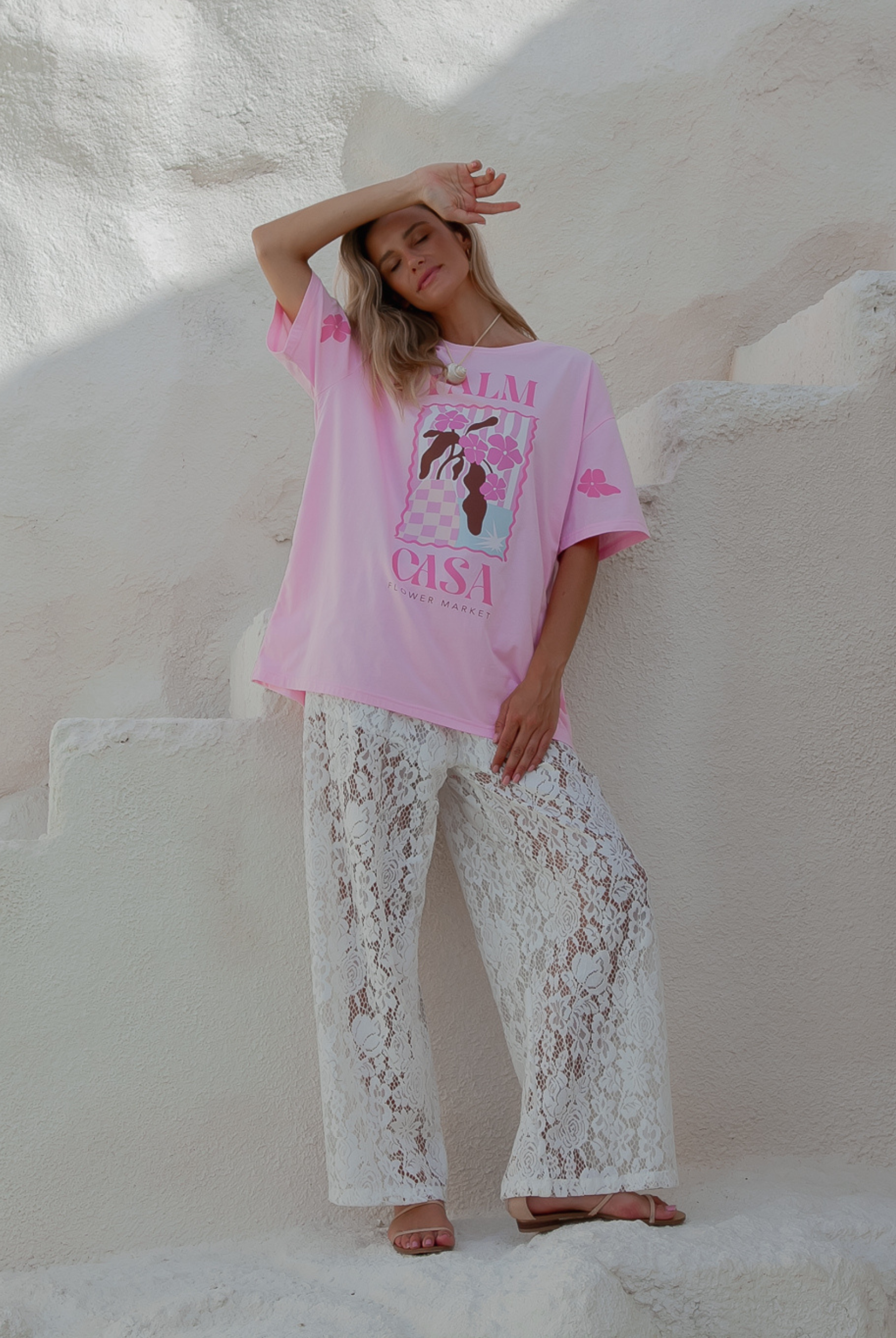 Person wearing a pink oversized t-shirt with text and graphics, paired with white lace pants, standing against a white stone wall.