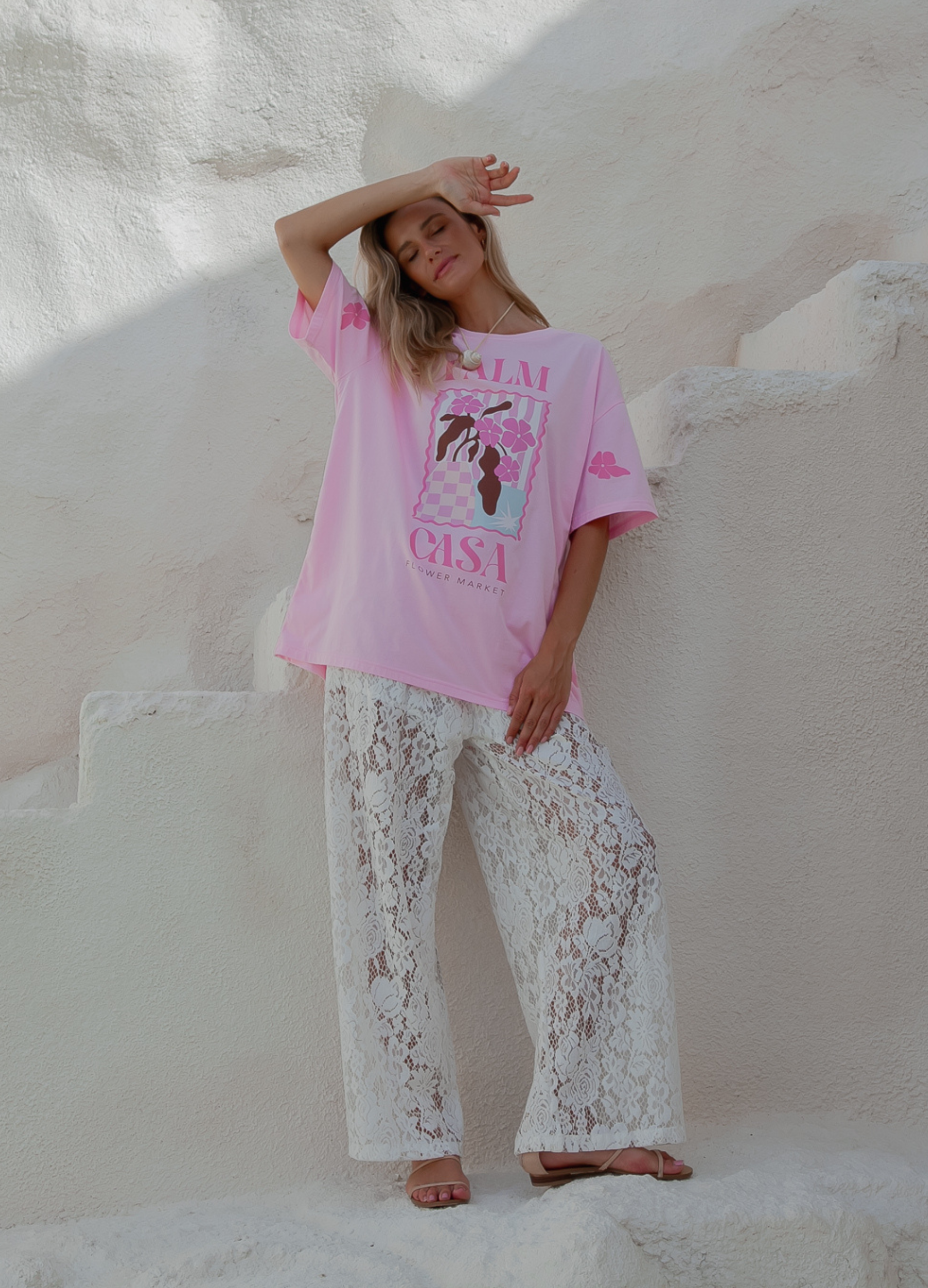 Person wearing a pink oversized t-shirt with text and graphics, paired with white lace pants, standing against a white stone wall.