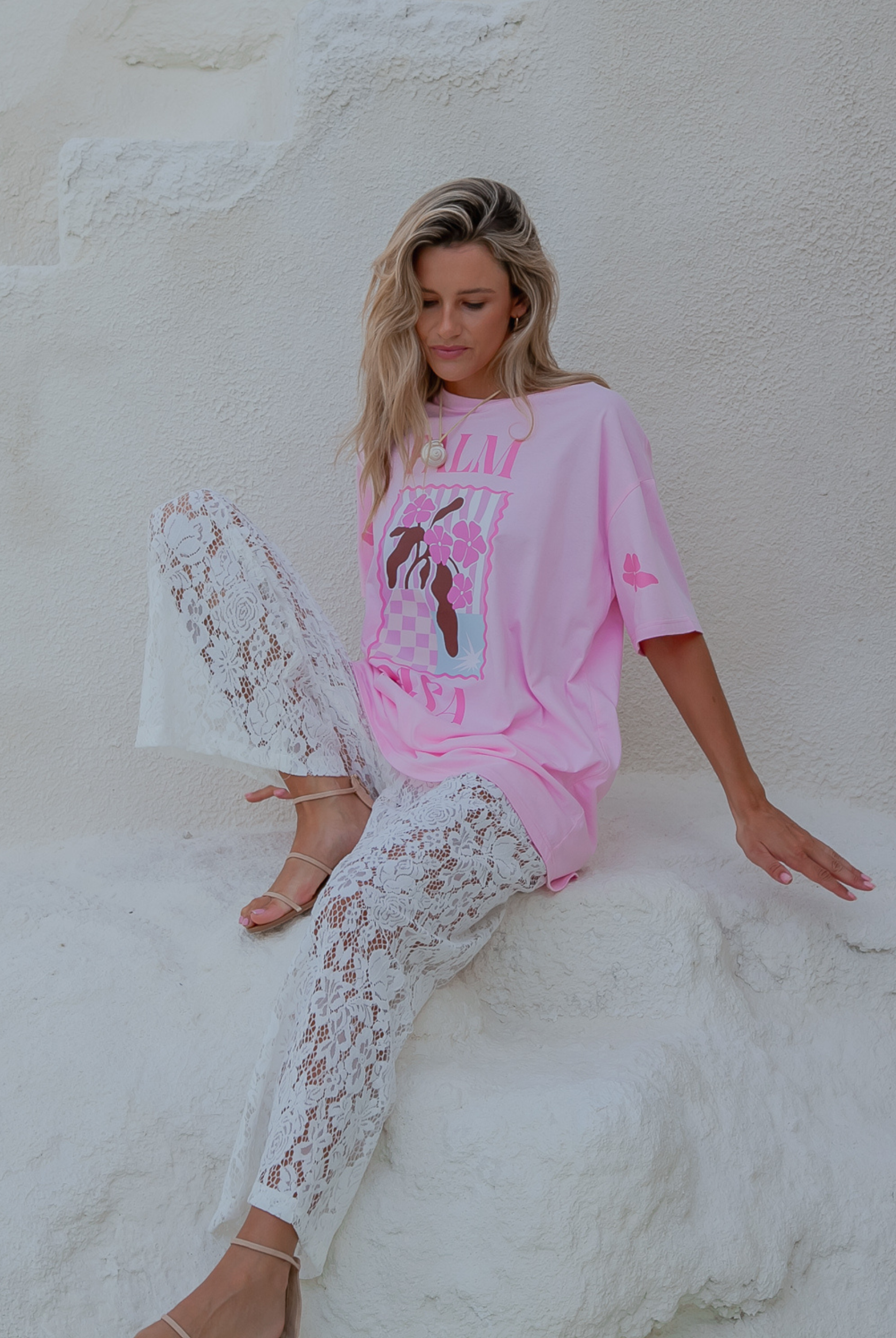 Woman wearing a pink oversized shirt and white lace pants sitting against a white wall.