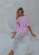 Woman wearing a pink oversized shirt and white lace pants sitting against a white wall.
