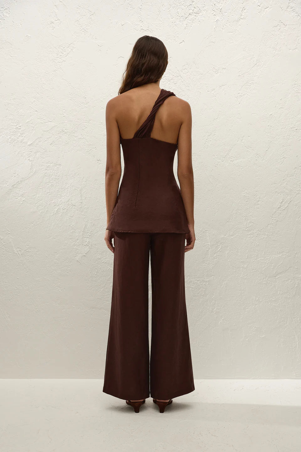 Person wearing a brown one-shoulder top and wide-leg pants against a white textured wall.