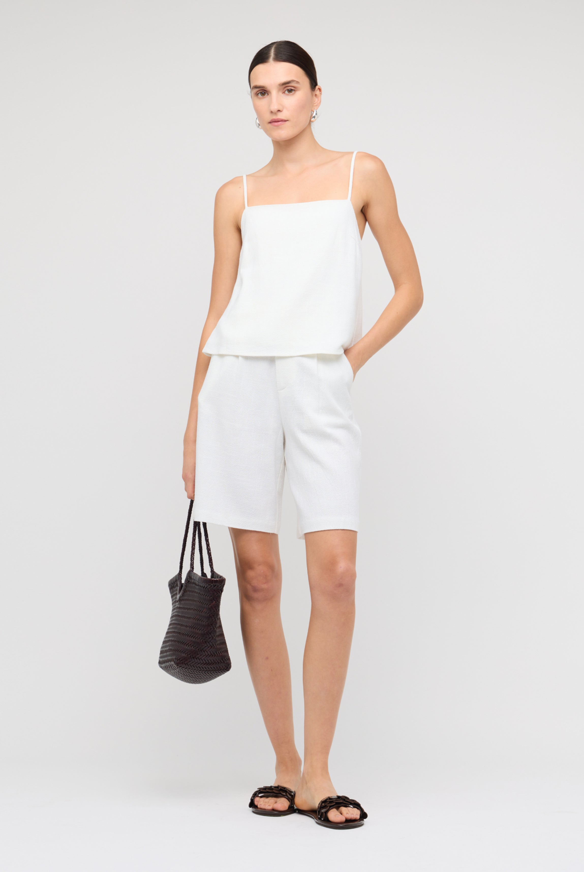 Woman wearing a white sleeveless top and shorts with a black handbag on a plain background