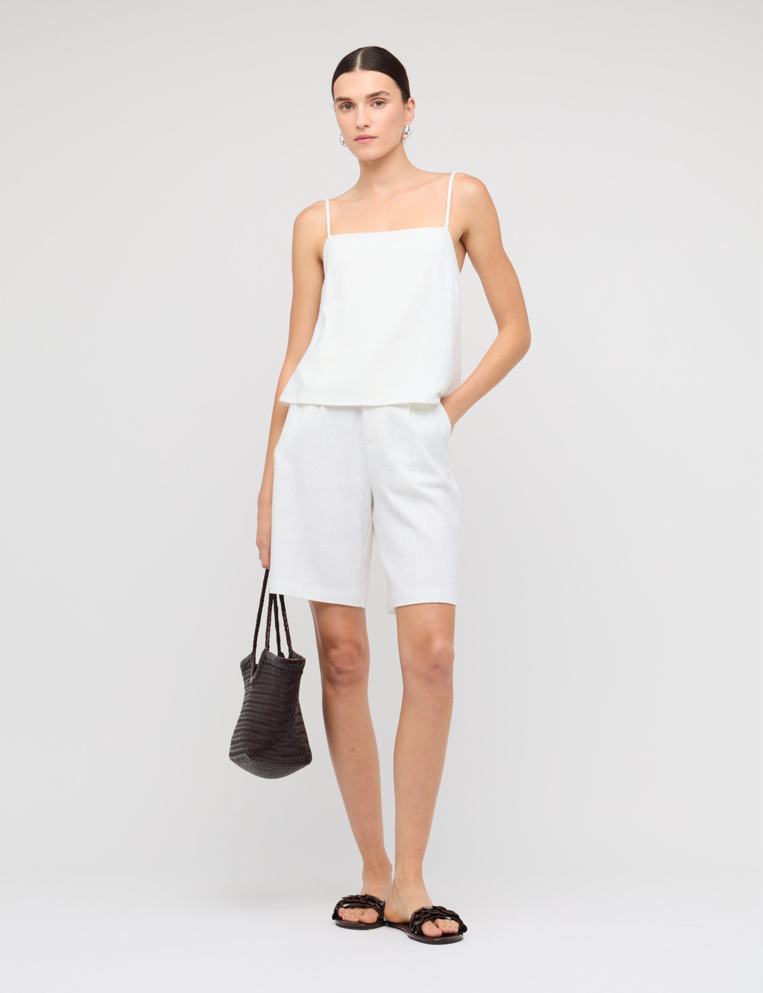 Woman wearing a white sleeveless top and shorts with a black handbag on a plain background