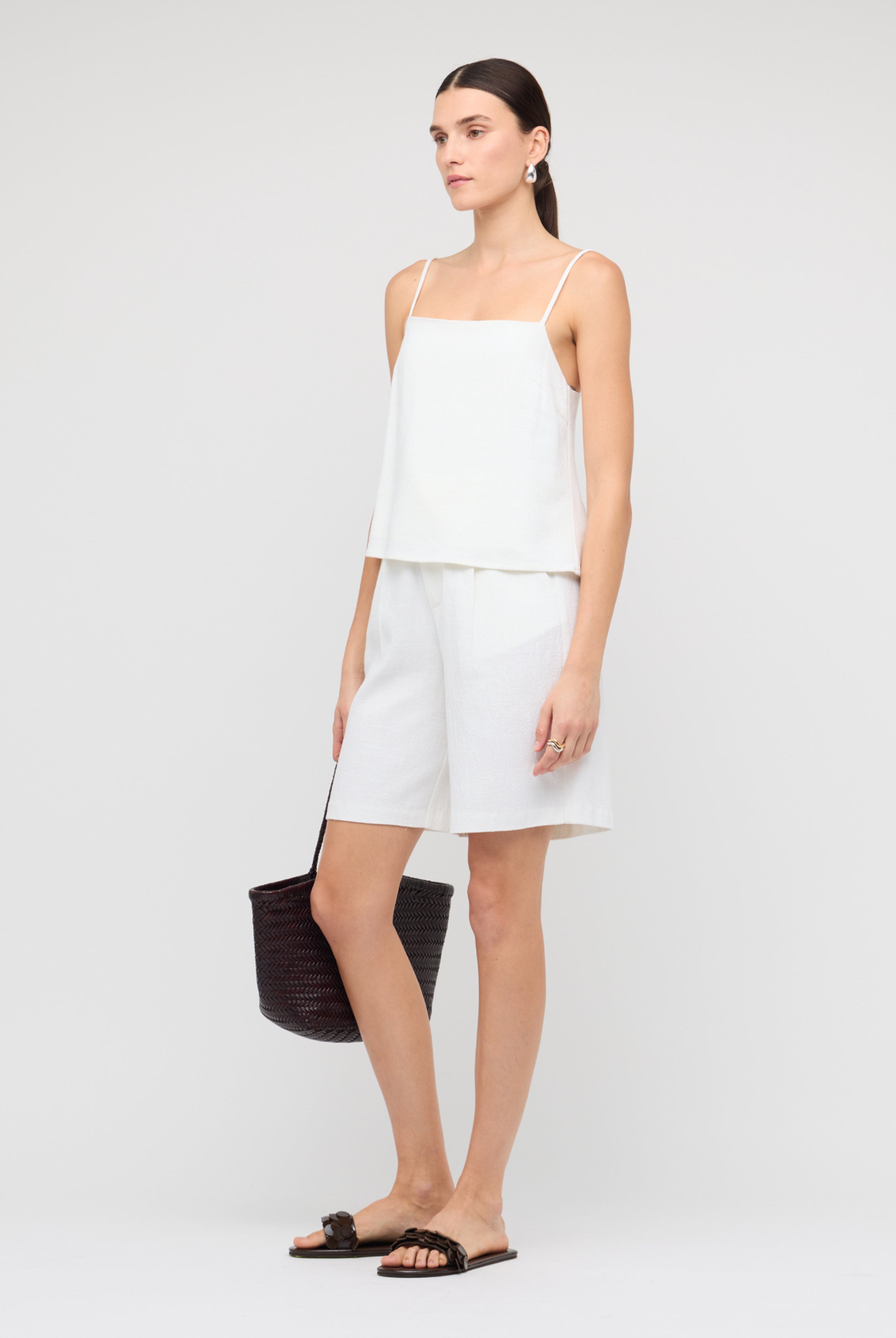 Woman wearing a white sleeveless top and shorts with a black handbag on a light gray background