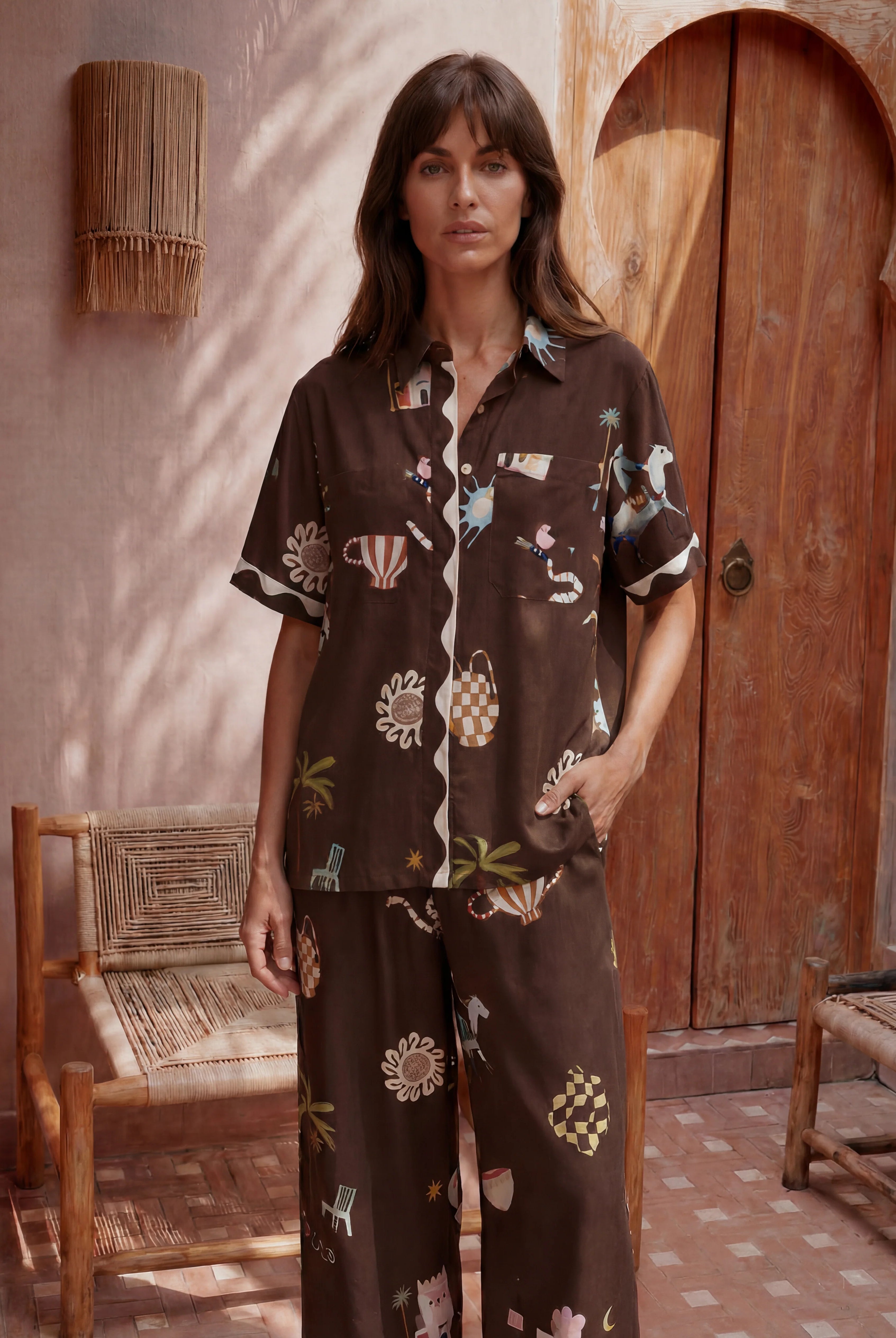 Woman wearing a patterned outfit standing in a sunlit room with wooden furniture.