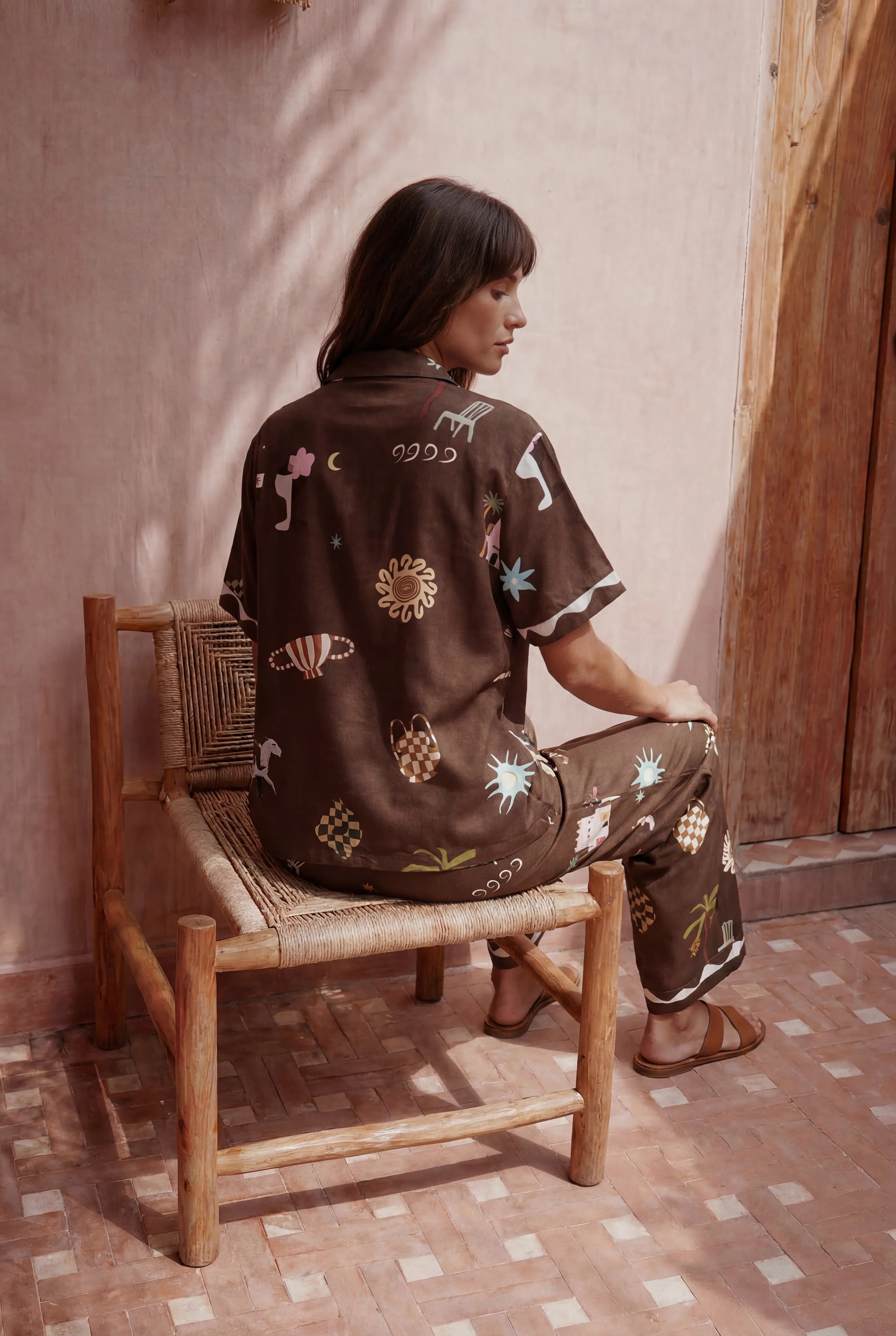 Woman sitting on a wooden chair wearing a patterned outfit in a warm-toned setting.