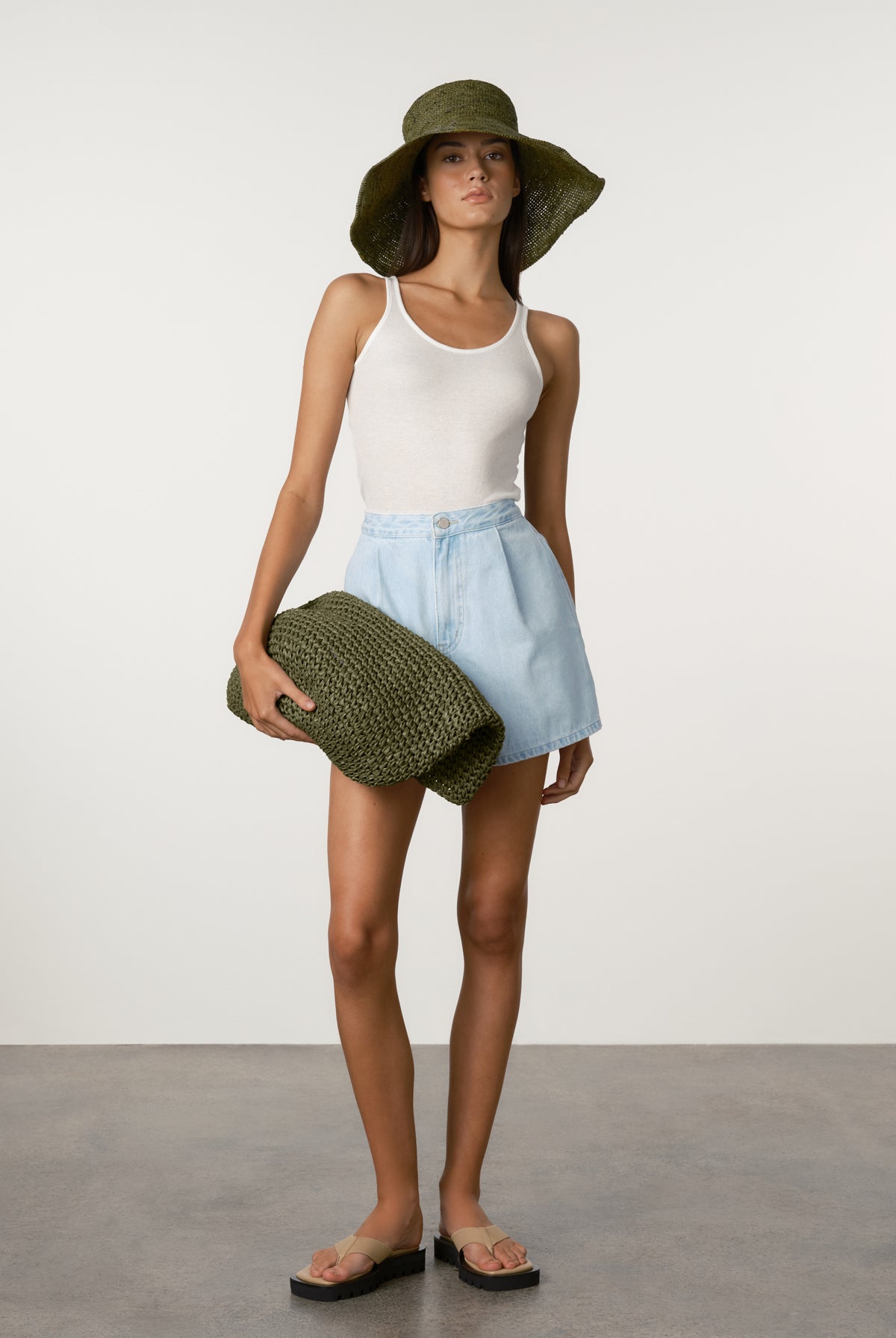Woman wearing a white tank top, light blue shorts, and a green hat holding a green clutch against a plain background.