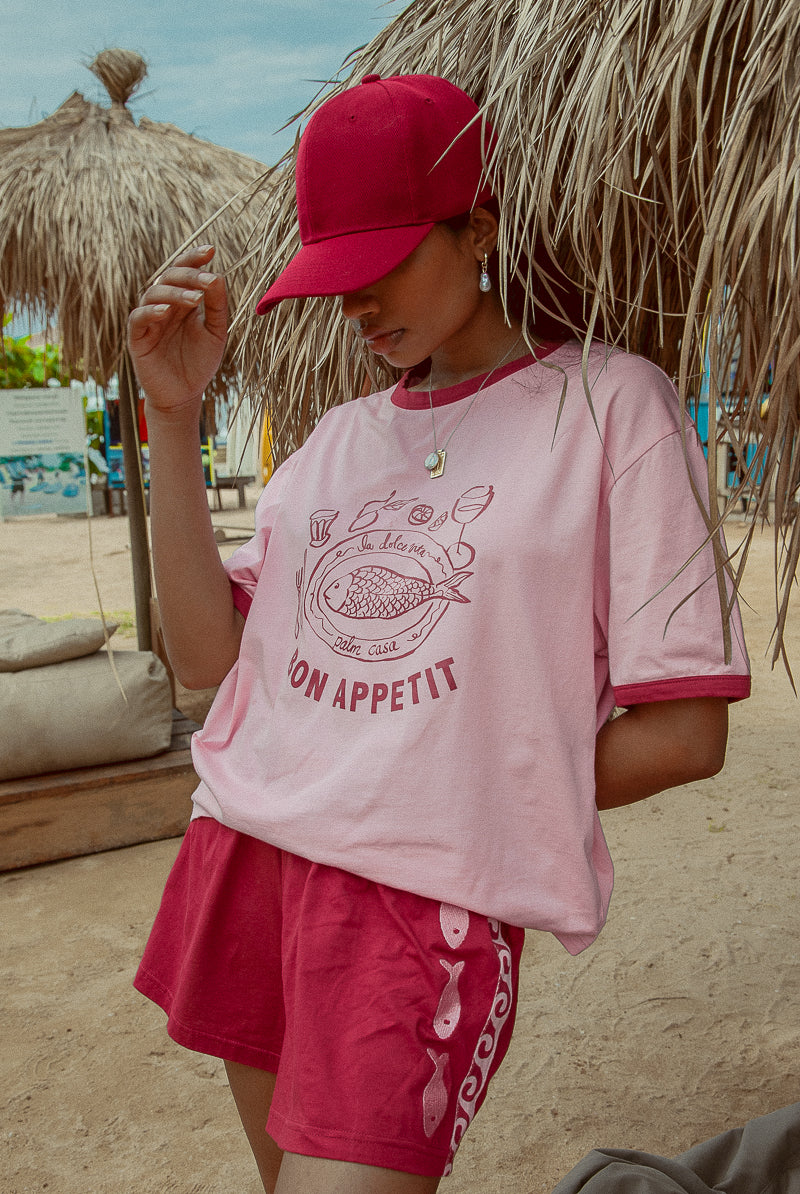 A person wearing a pink 'Bon Appetit' graphic tee with maroon binding on the neck and sleeves, paired with matching shorts, standing in a beach setting.