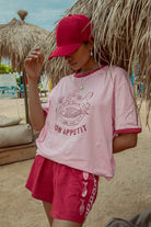 A person wearing a pink 'Bon Appetit' graphic tee with maroon binding on the neck and sleeves, paired with matching shorts, standing in a beach setting.
