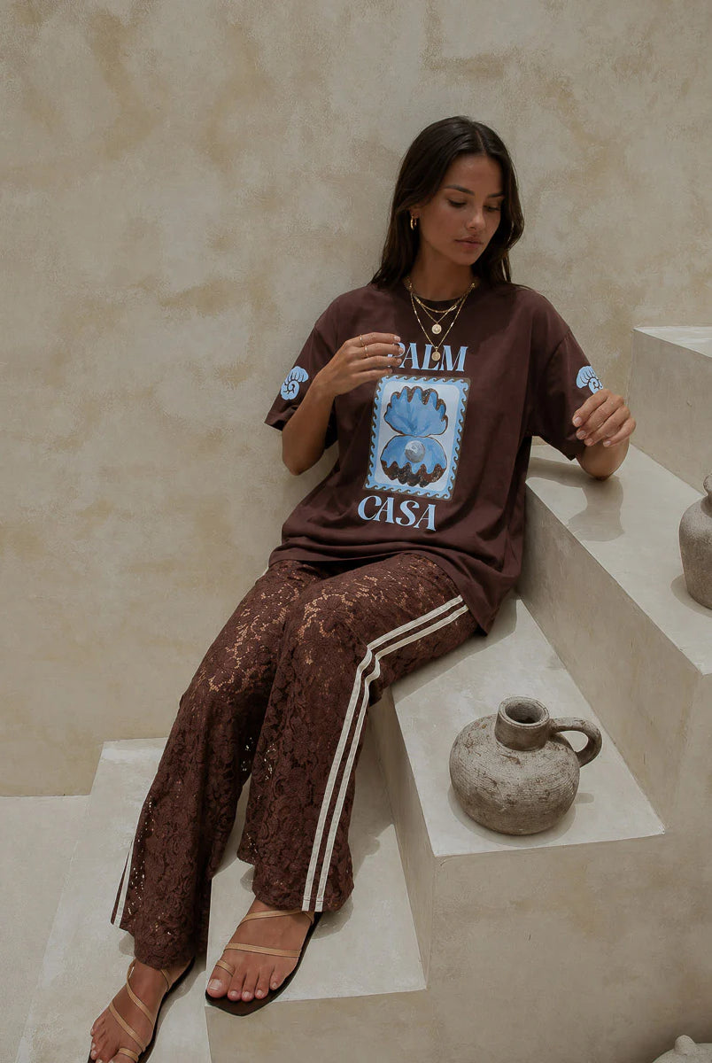 Woman sitting on steps wearing a brown graphic t-shirt and pants, surrounded by decorative items.