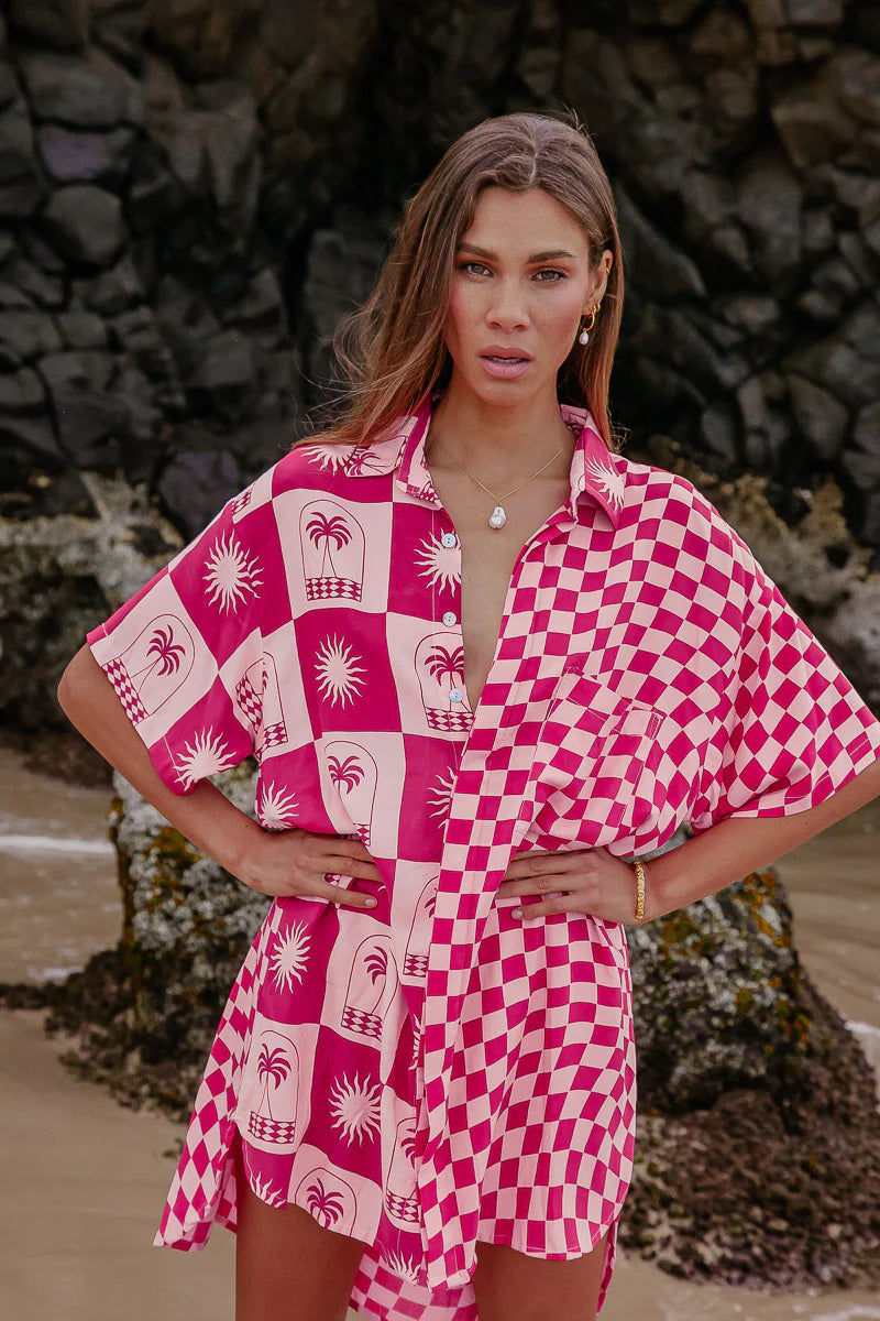 Woman wearing a pink and white patterned shirt with a checkered design, standing against a natural background.