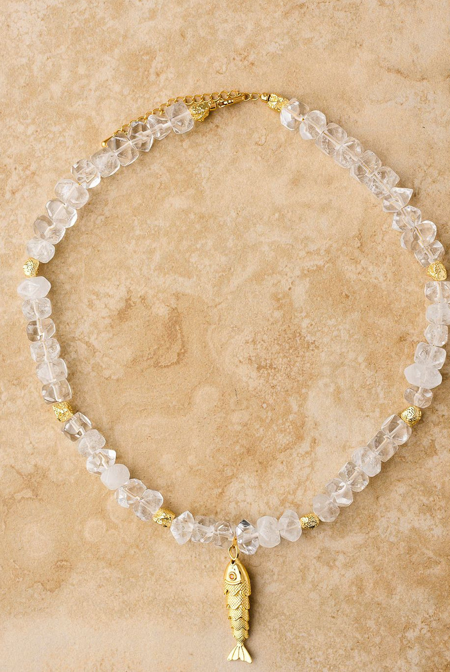 Necklace with clear beads and gold fish charm on a beige stone surface