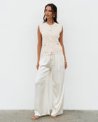 A woman wearing a sleeveless cream-colored vest with metal buttons, paired with white trousers. Accessory jewelry is also visible.