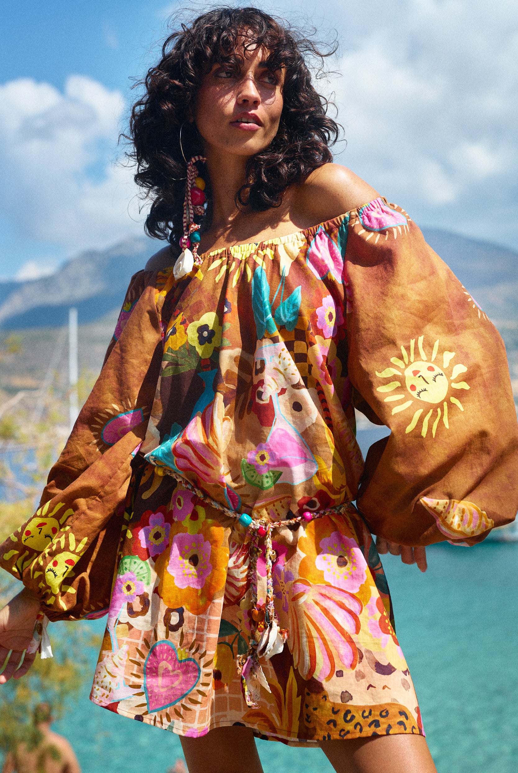 Woman wearing a colorful off-shoulder dress with floral patterns and a sun design, standing outdoors with mountains in the background.