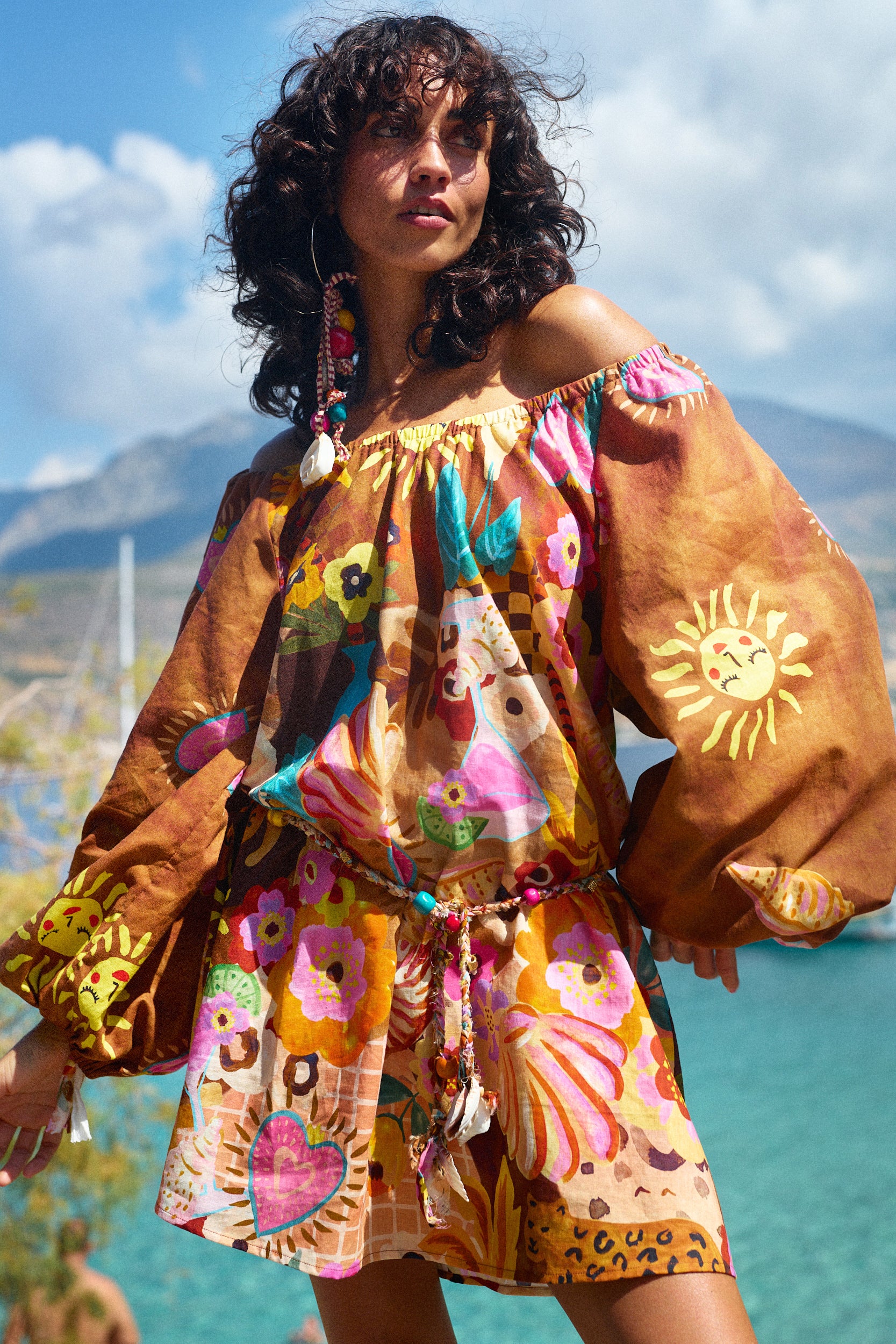 Woman wearing a colorful off-shoulder dress with floral patterns and a sun design, standing outdoors with mountains in the background.
