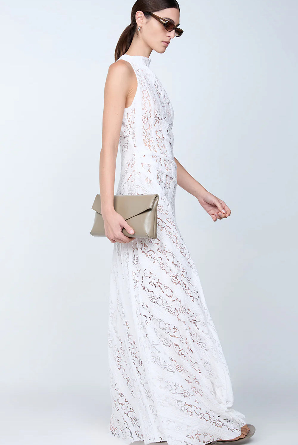 Woman in a white lace dress holding a beige clutch against a white background