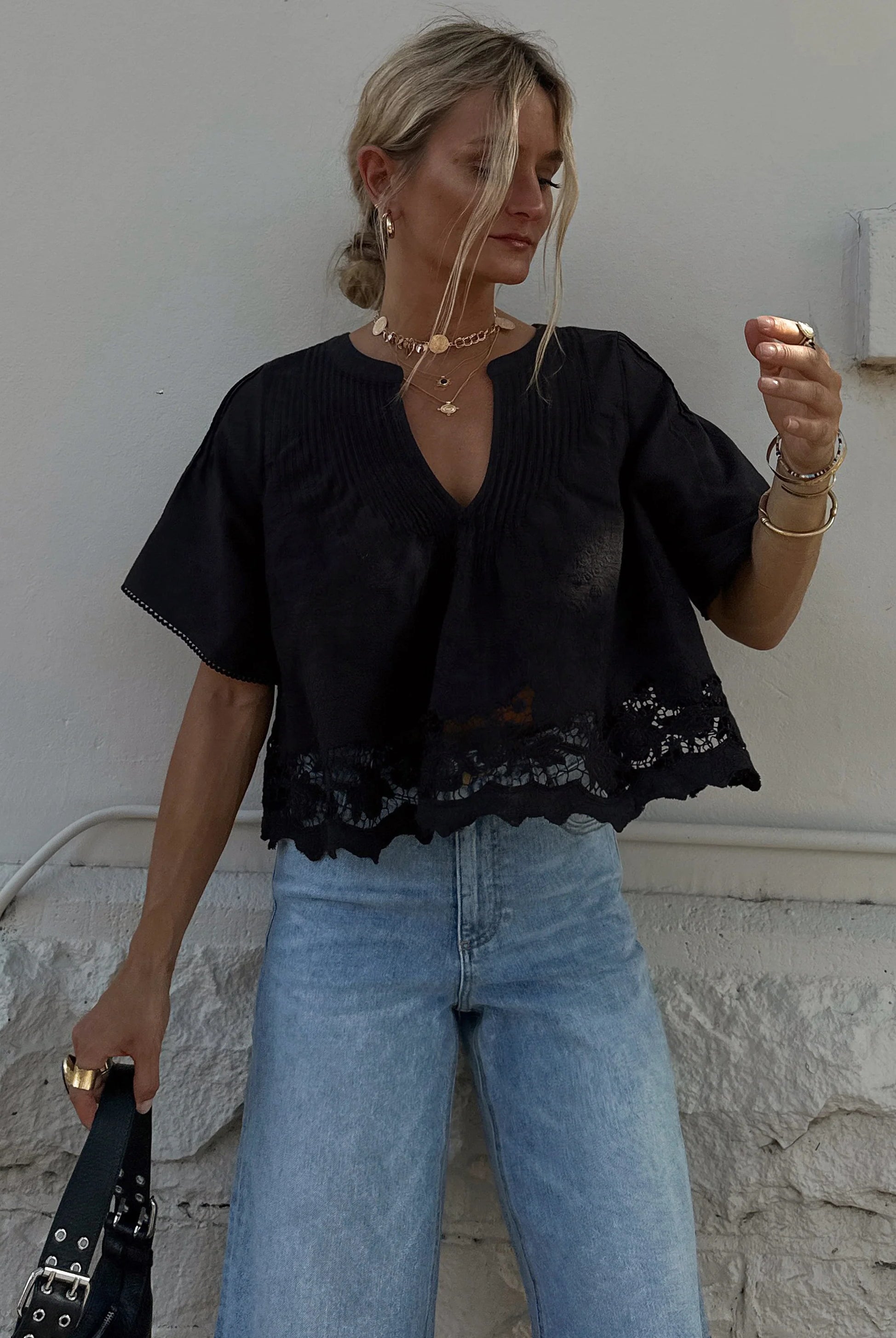 Woman wearing a black top with lace details and blue jeans against a white wall.