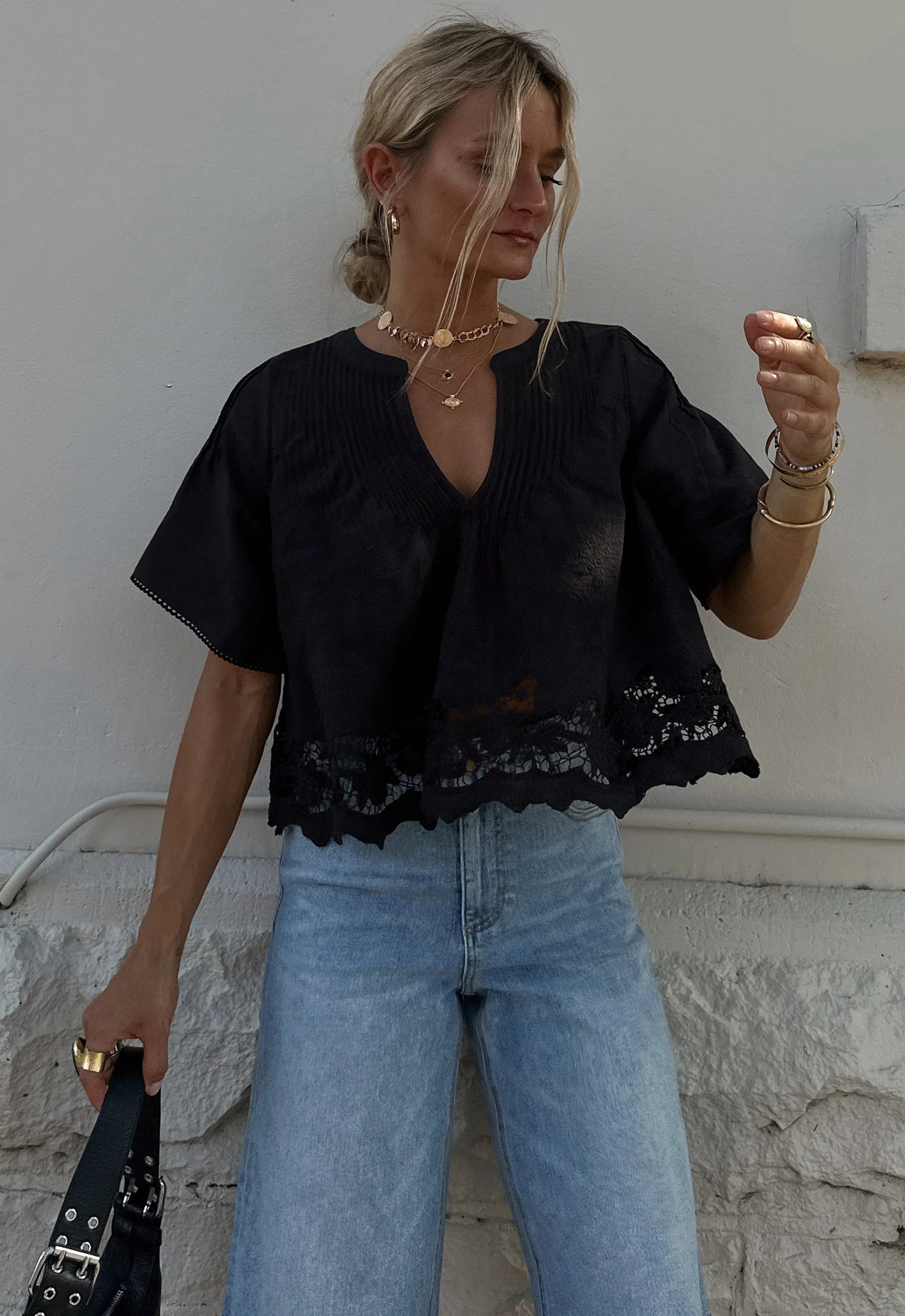 Woman wearing a black top with lace details and blue jeans against a white wall.