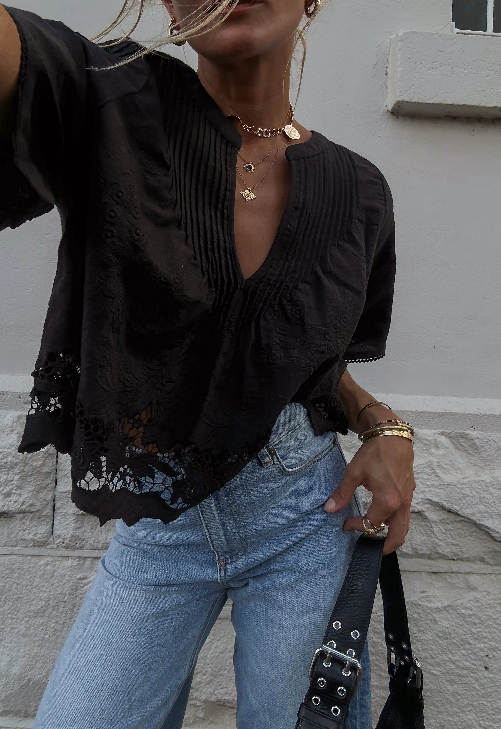 Person wearing a black top with lace details and blue jeans, standing against a light gray wall.