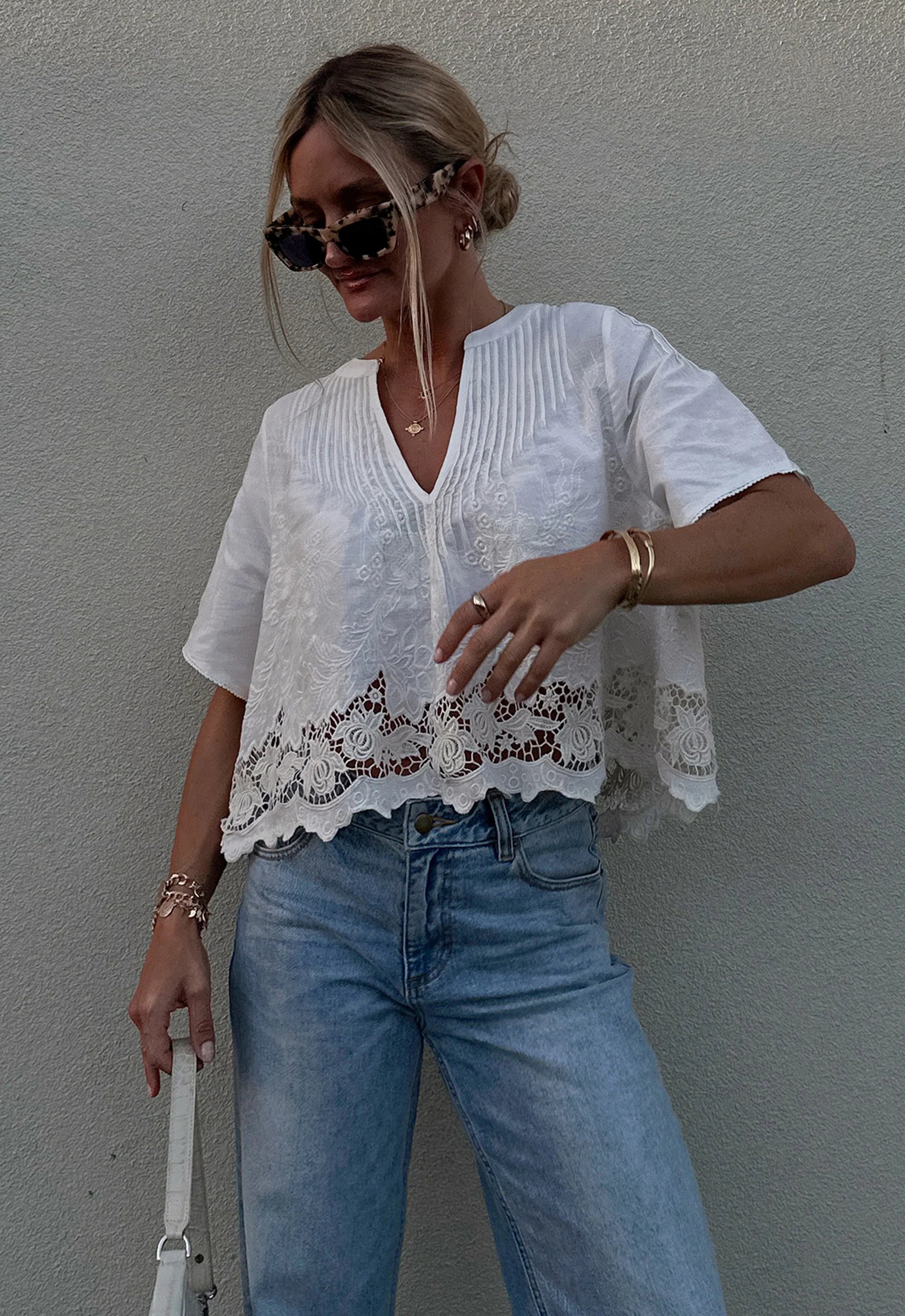 Woman wearing a white lace top and blue jeans against a gray wall