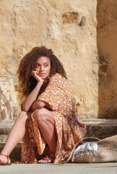 Woman sitting on a stone wall with a floral shawl draped over her, against a rustic stone wall background.