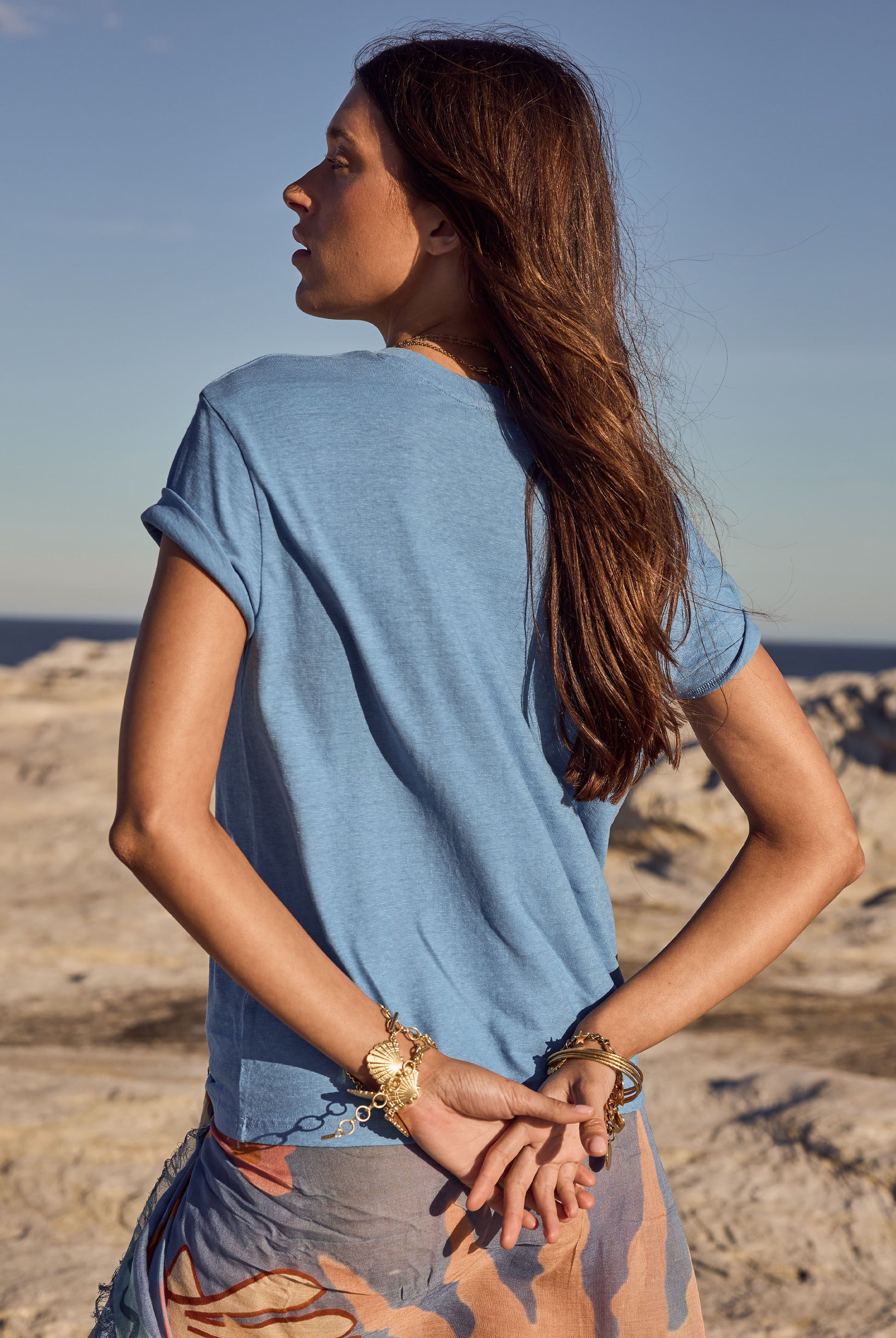 Woman in a blue shirt standing on a rocky beach with a clear sky.