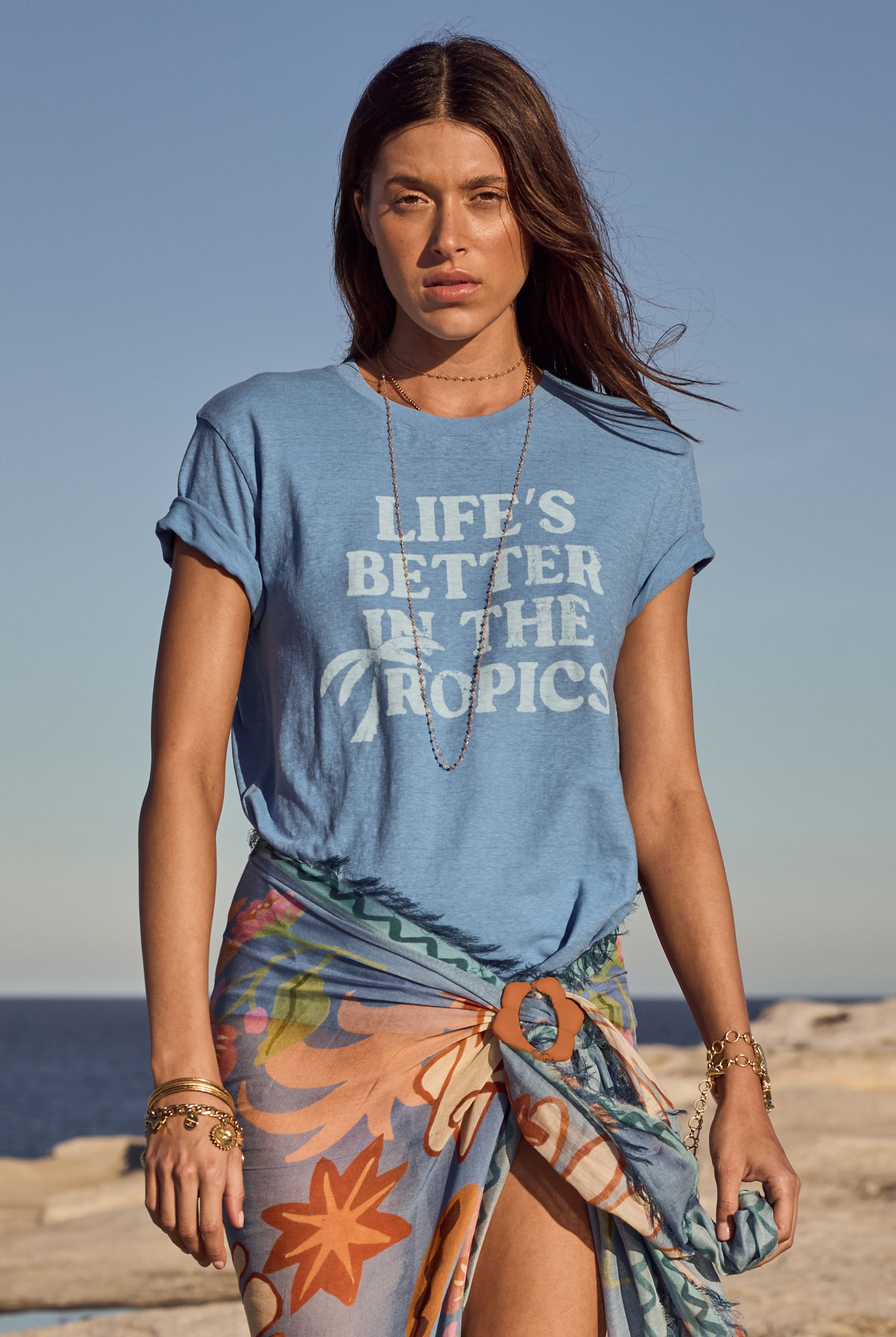Woman wearing a blue t-shirt with 'Life's Better in the Tropics' text on a beach.