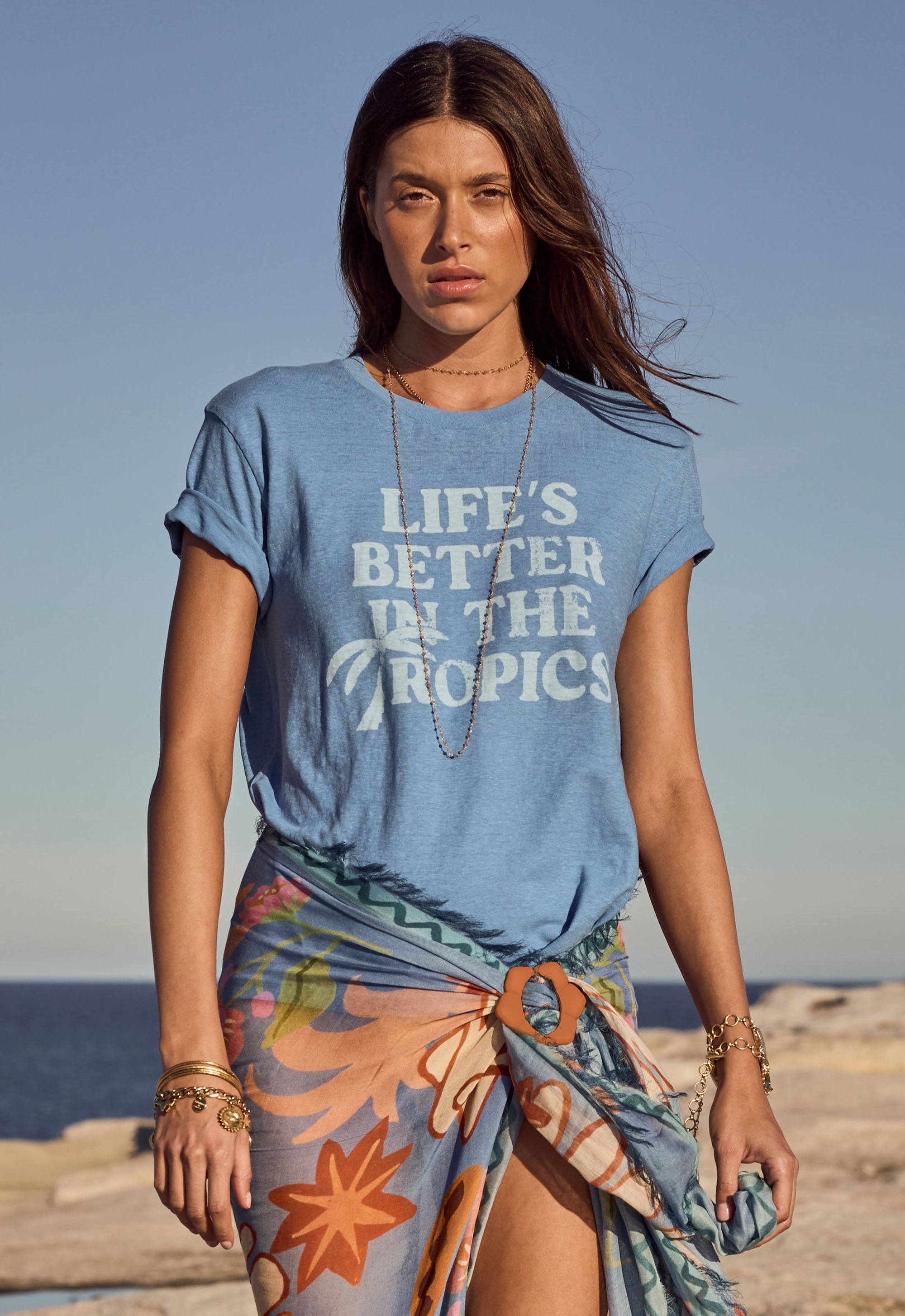 Woman wearing a blue t-shirt with 'Life's Better in the Tropics' text on a beach.