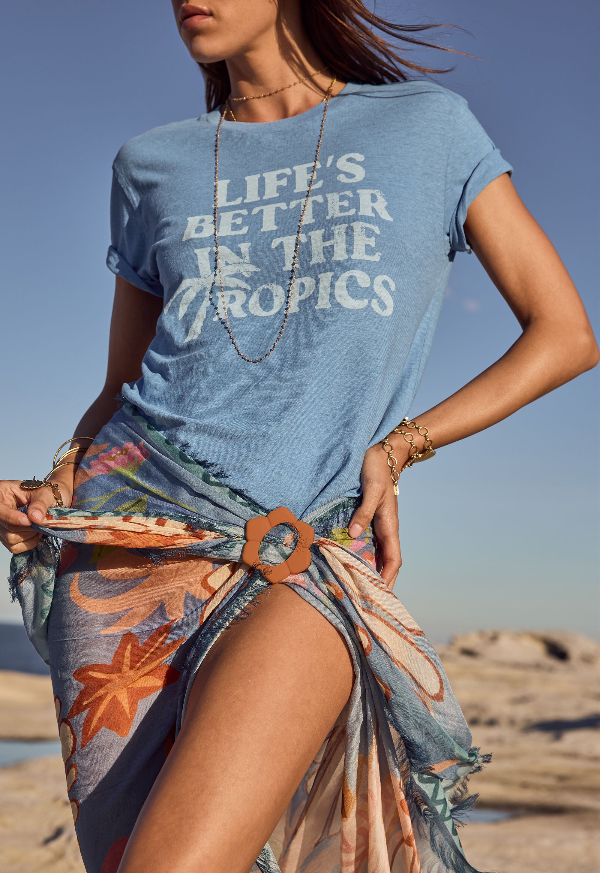 Person wearing a blue t-shirt with 'Life's Better in the Tropics' text on a beach.