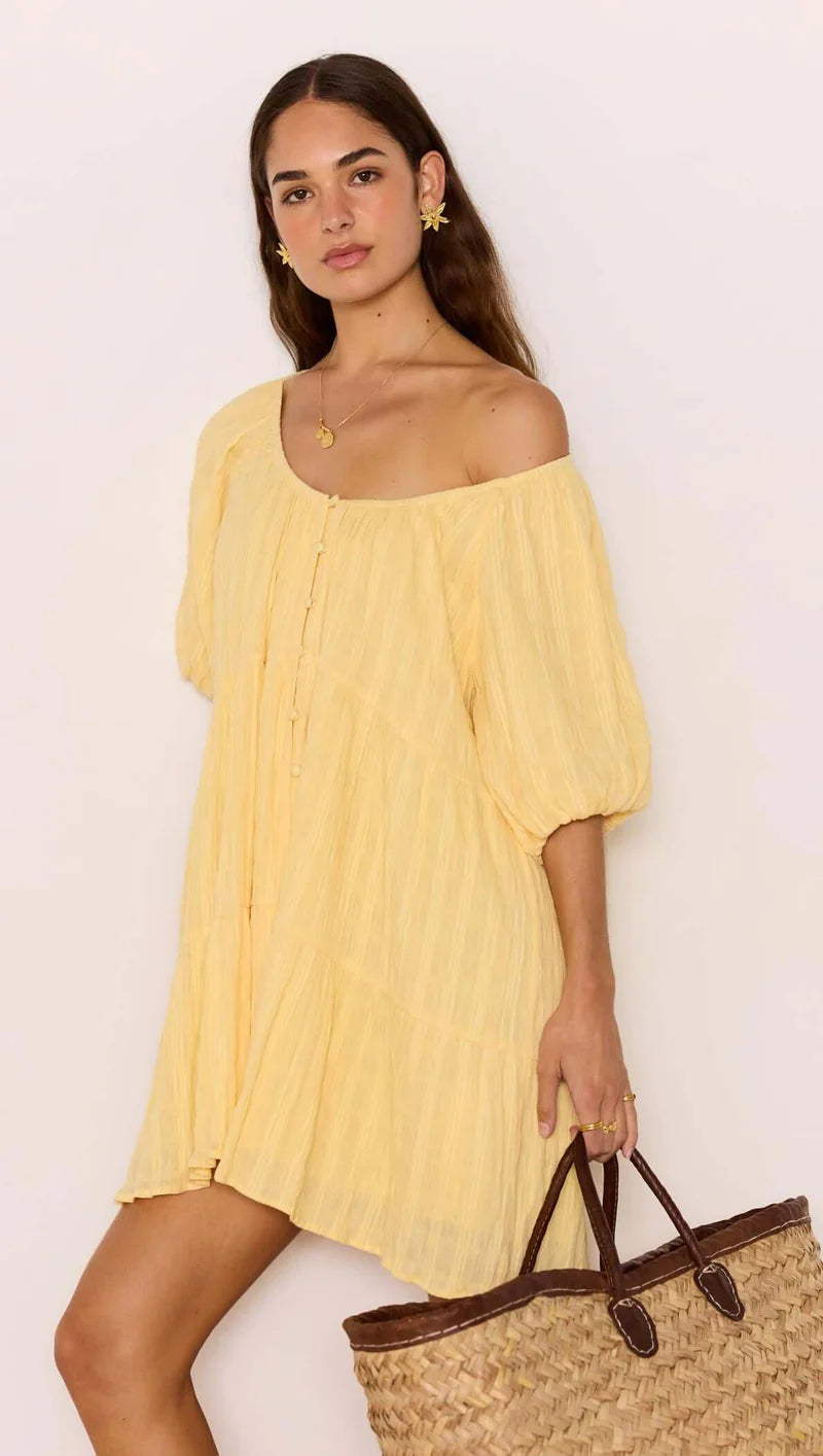 Woman wearing a yellow off-shoulder dress holding a straw bag against a white background