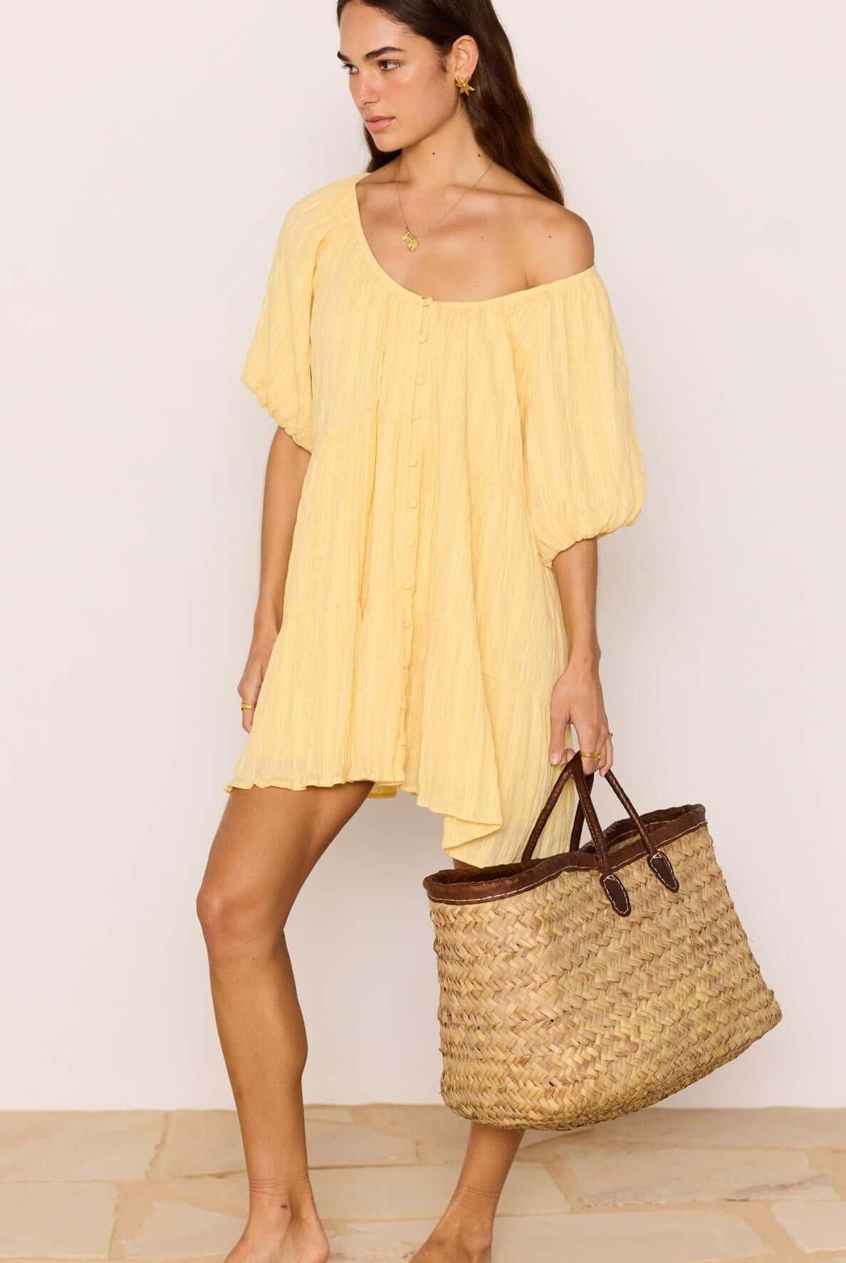 Woman in a yellow dress holding a straw bag against a plain background