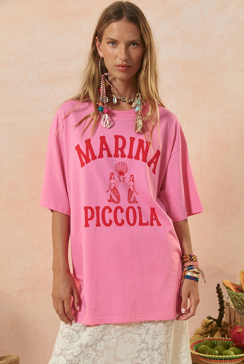 A woman wearing a pink oversized t-shirt with 'MARINA PICCOLA' printed in red text on the front. She is also accessorized with multiple necklaces and bracelets and is standing against a pink background.