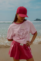 A person wearing a pink 'Bon Appetit' graphic tee with maroon binding on the neck and sleeves, paired with matching shorts, standing in a beach setting.
