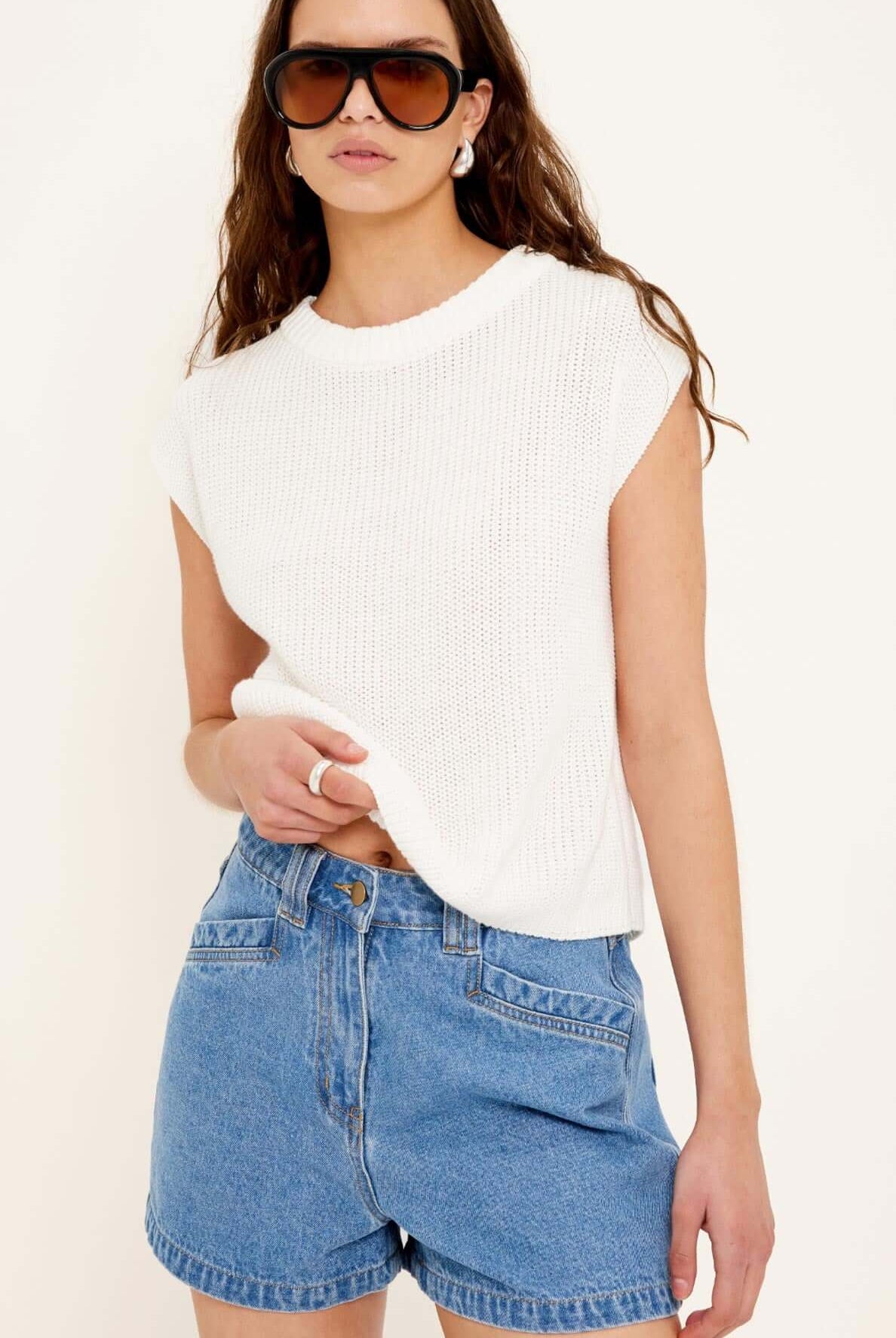 Woman wearing a white sleeveless top and blue denim shorts on a plain background