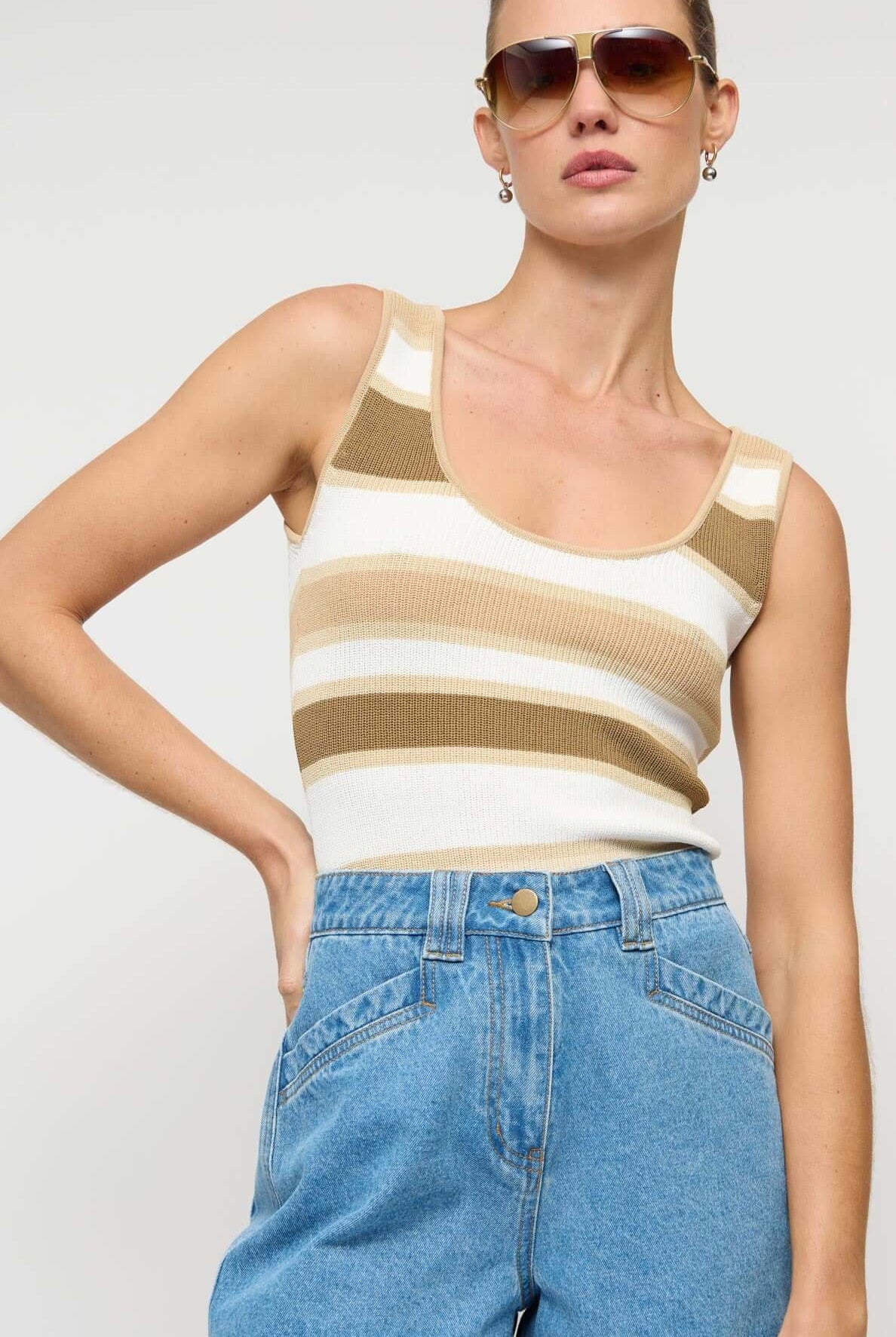Woman wearing a striped tank top and denim shorts with sunglasses on a plain background
