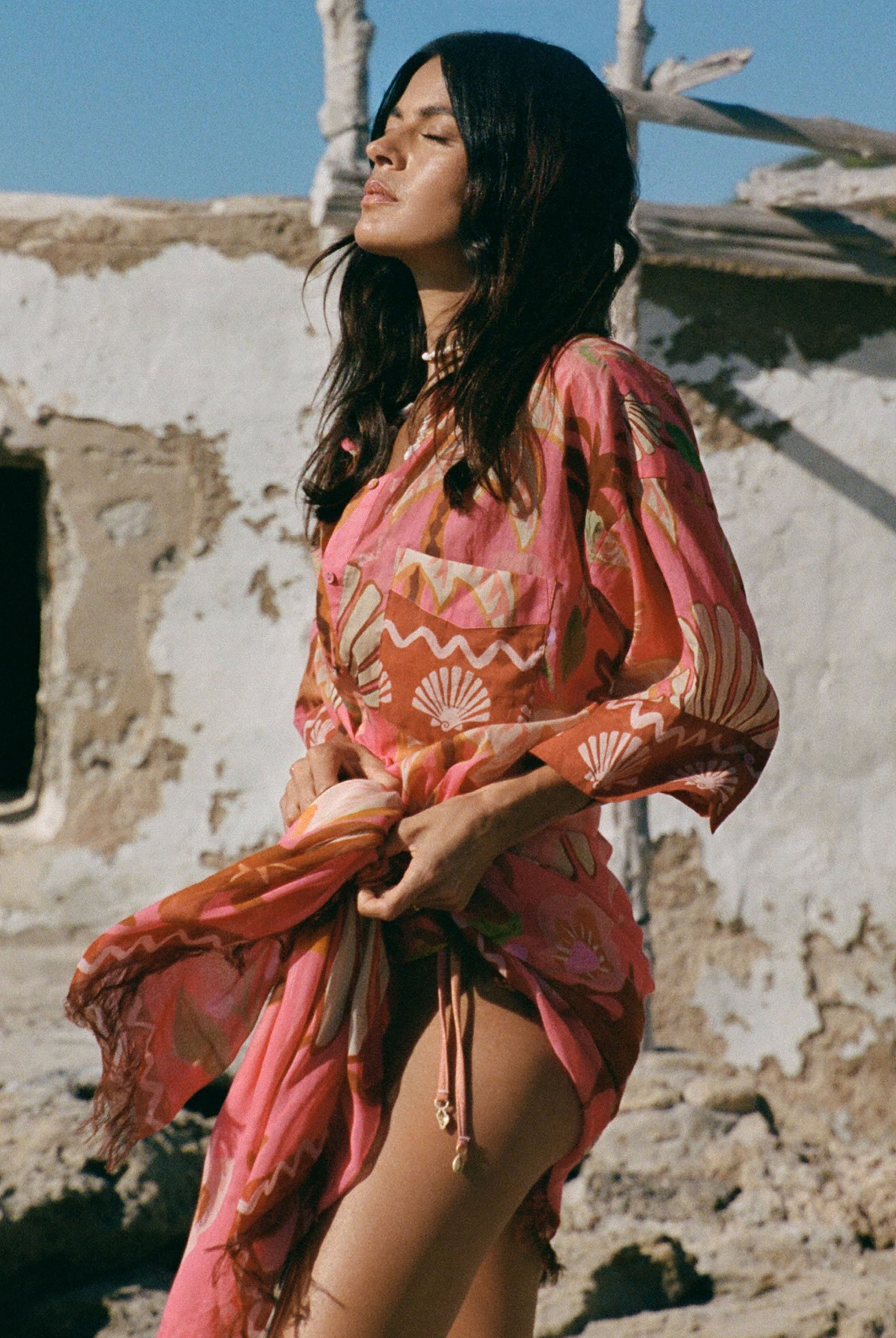 Woman in a pink and orange patterned dress standing in front of a rustic wall.
