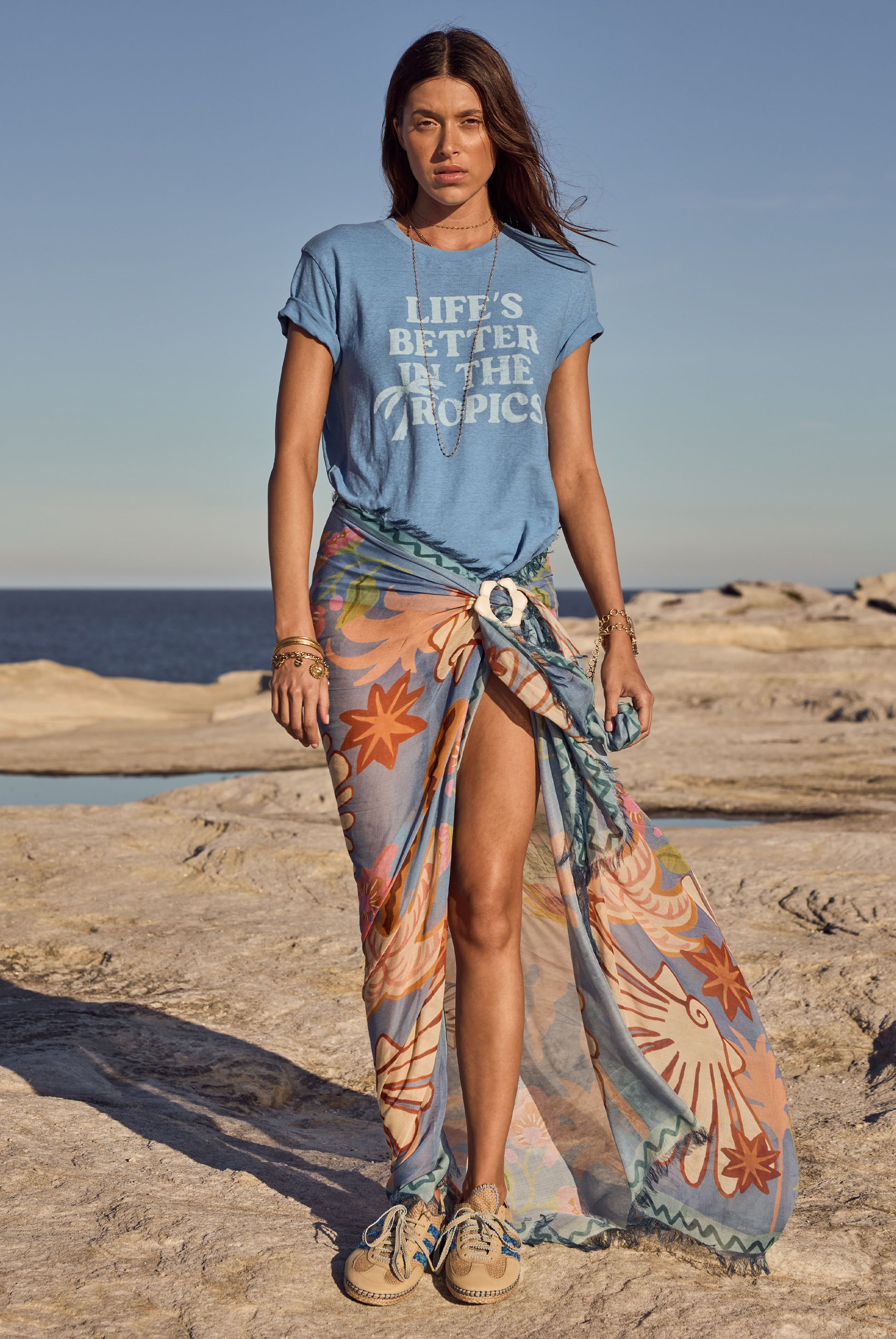 Woman on a beach wearing a blue t-shirt with 'Life's Better in the Tropics' text and a colorful sarong.