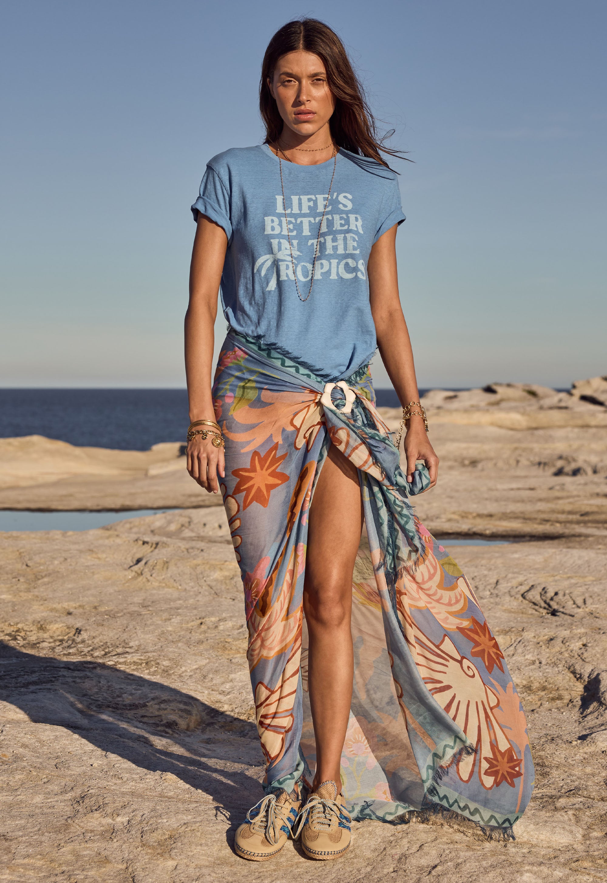 Woman on a beach wearing a blue t-shirt with 'Life's Better in the Tropics' text and a colorful sarong.