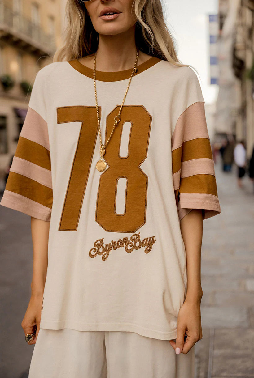 Person wearing a beige t-shirt with '78 Byron Say' print on a city street.
