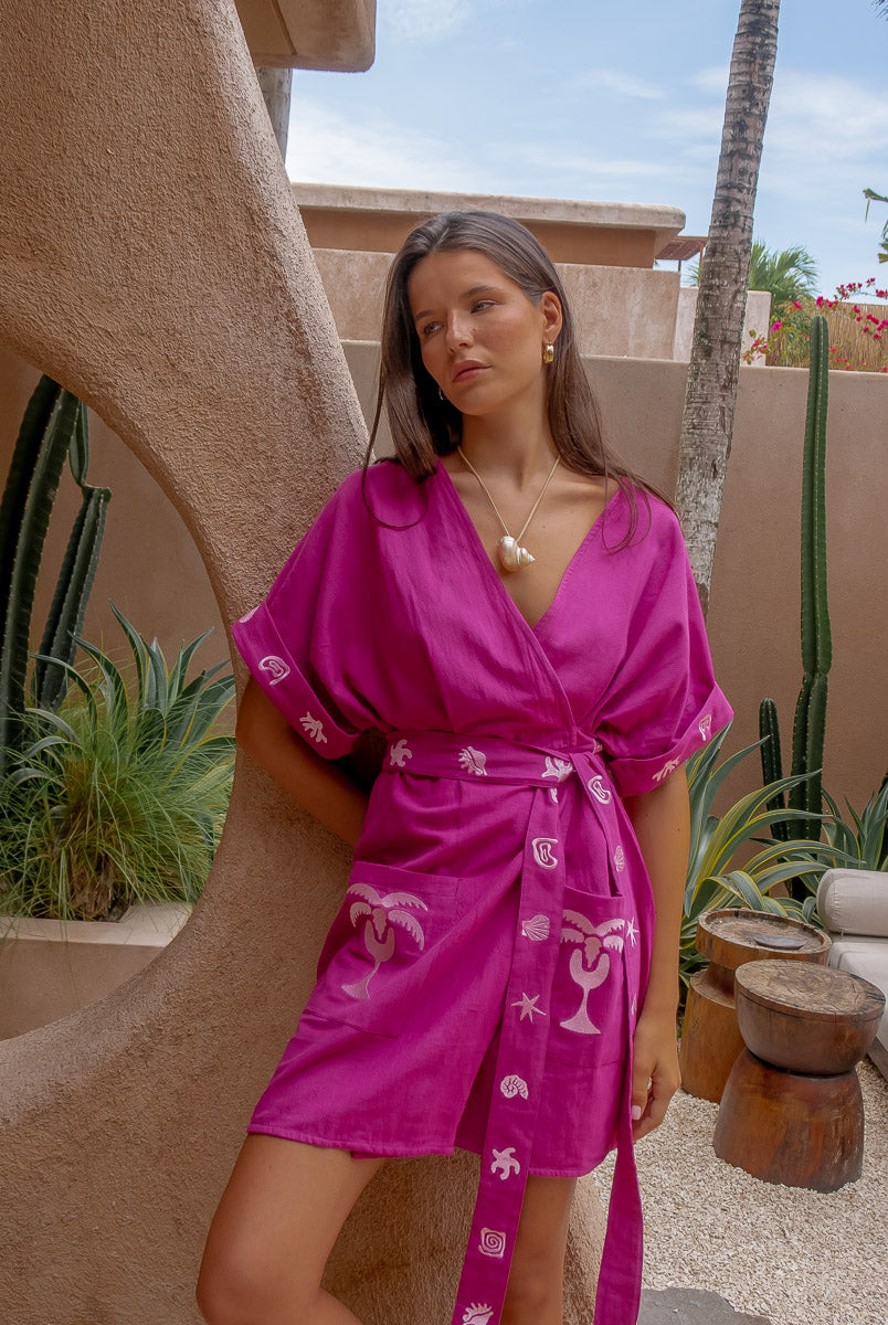 Woman wearing a pink dress with white patterns in an outdoor setting with plants and a building.
