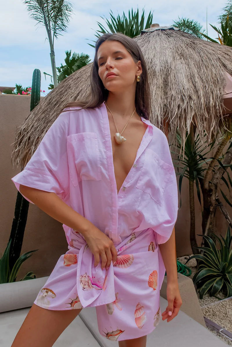 Woman wearing a pink robe with shell patterns under a thatched umbrella.
