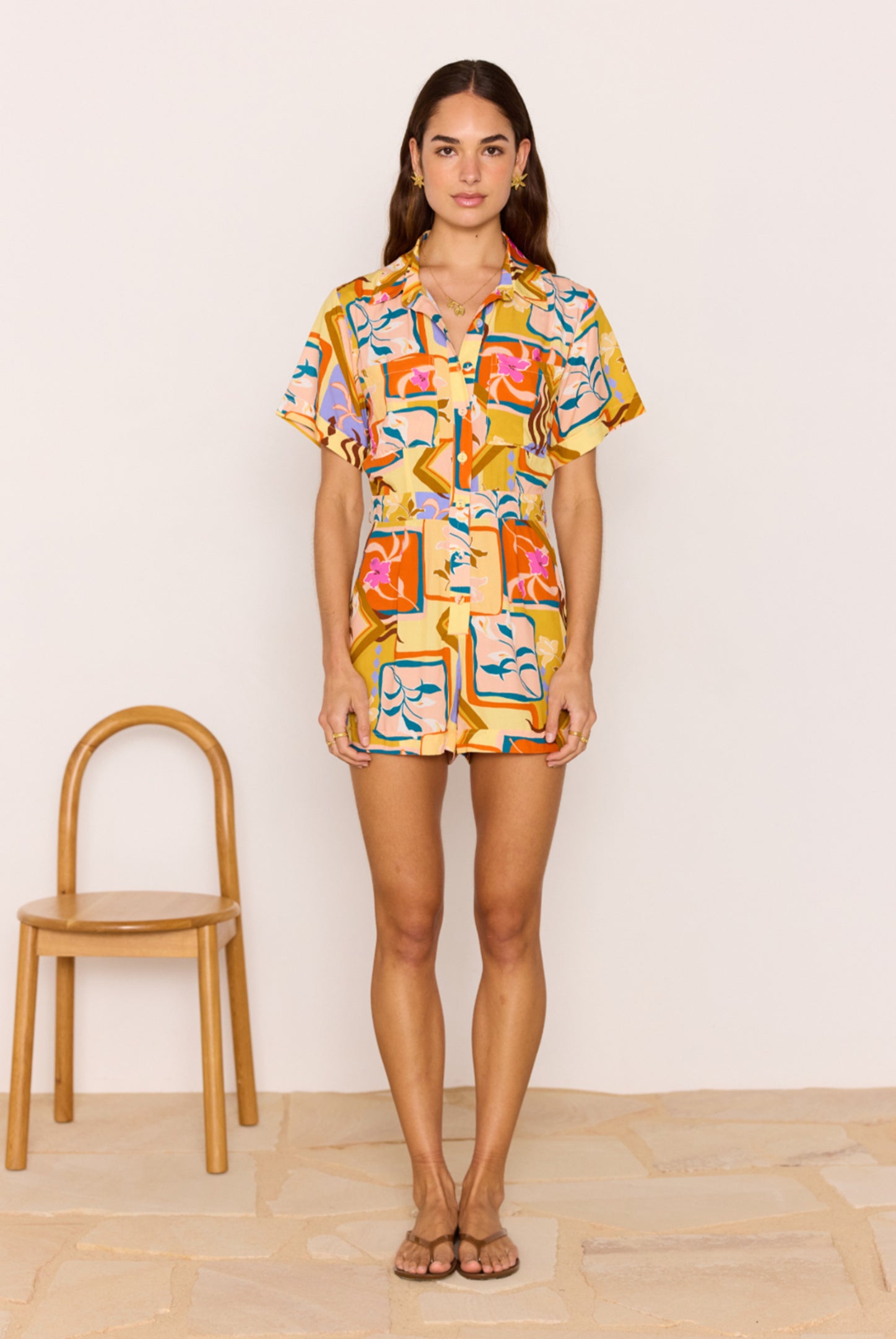 Woman wearing a colorful patterned outfit standing next to a wooden chair on a light wood floor.