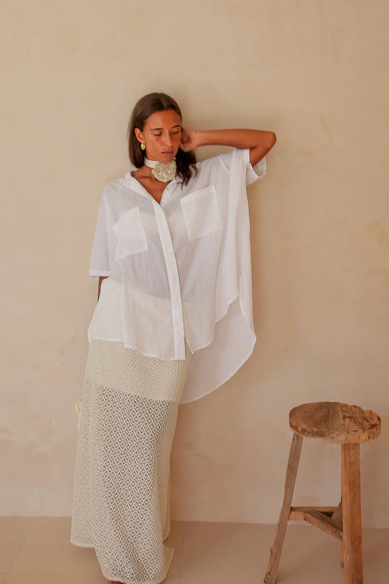 Woman in a white outfit standing next to a wooden stool against a beige wall.