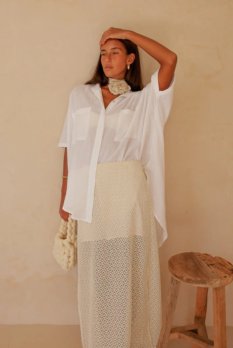 Woman wearing a white shirt and beige skirt against a beige wall with a wooden stool.