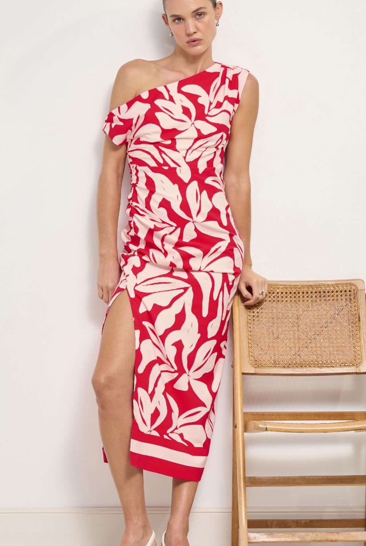 Woman wearing a red and white patterned dress standing next to a wooden chair against a white wall.