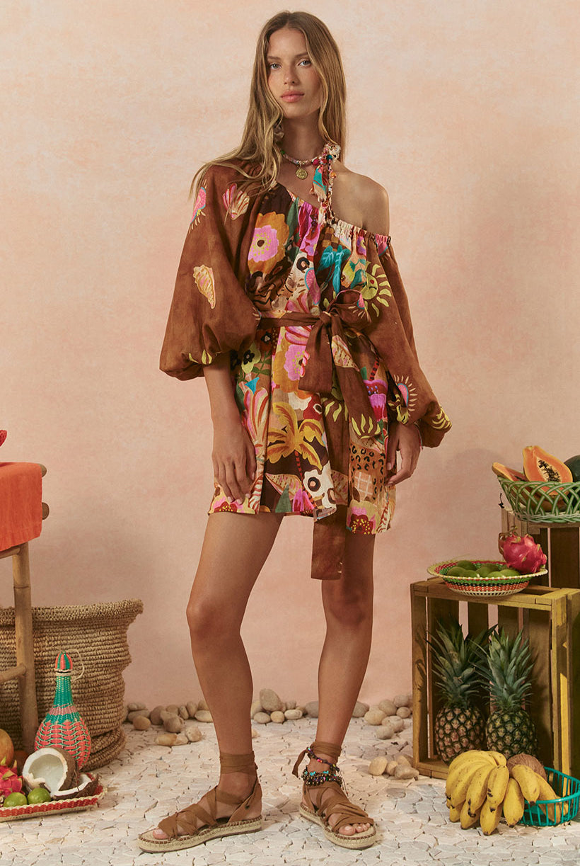 A model wearing a sleeveless tunic dress with a floral pattern in a tan color, standing in a setting with tropical fruits and decorative items.