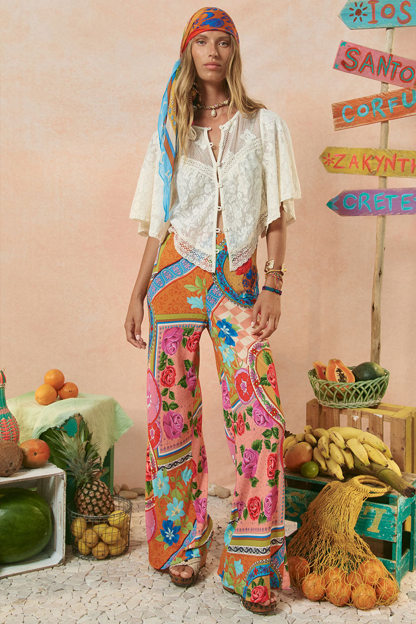 Person wearing a colorful outfit with floral pants and a headscarf, standing in front of fruit and decorative signs.