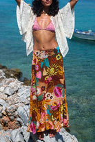 Woman in a colorful skirt and pink bikini top standing on rocks by the ocean.