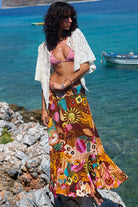 Woman in a colorful skirt and bikini top standing on rocks by the ocean with boats in the background.