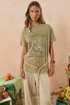 Woman wearing a green graphic t-shirt with a truck design, standing in a casual setting with fruits and a pink background.